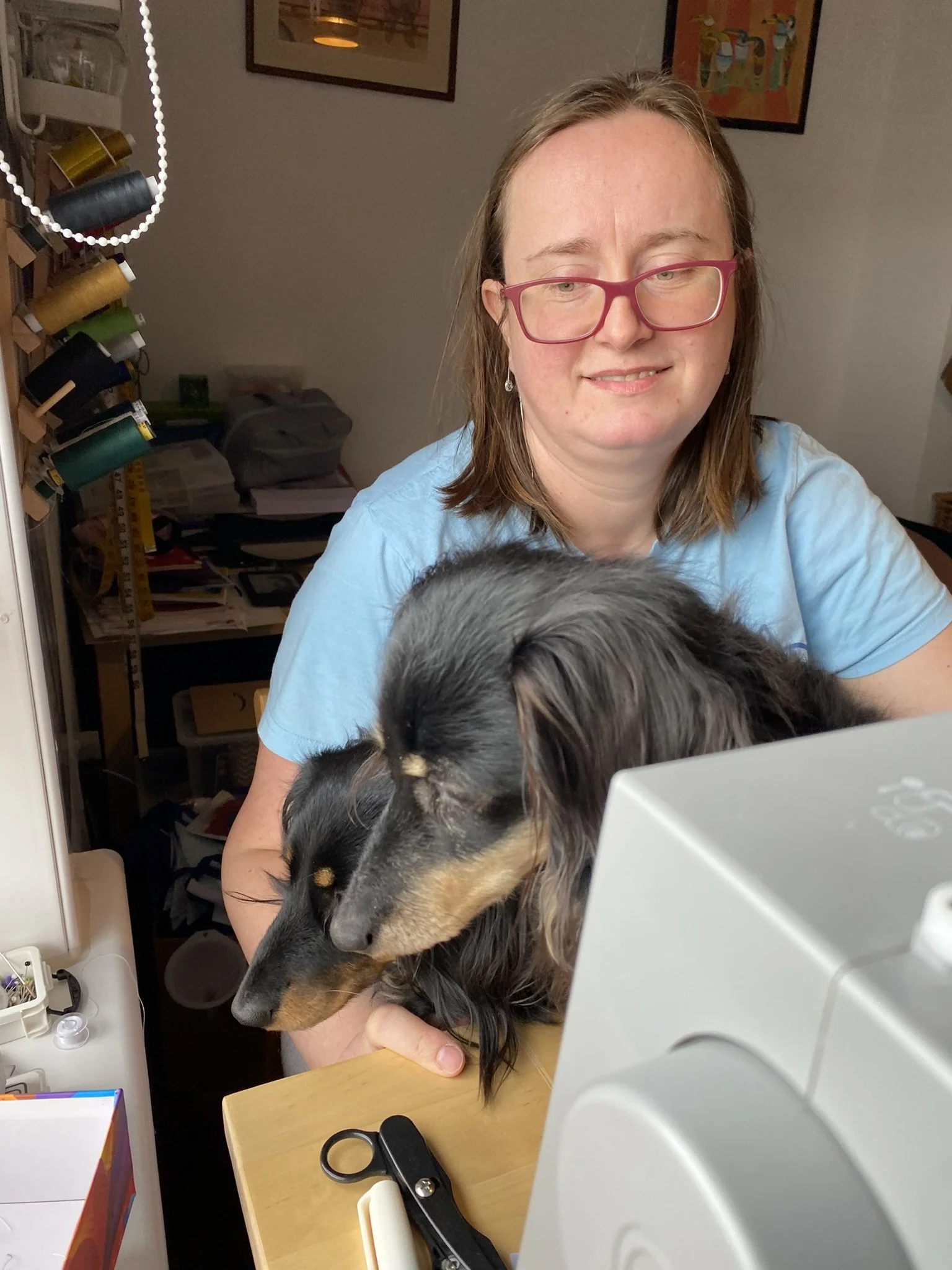 A woman with glasses and long hair sitting at a desk with two black and tan dogs on her lap, looking down at a computer monitor in a cluttered room.