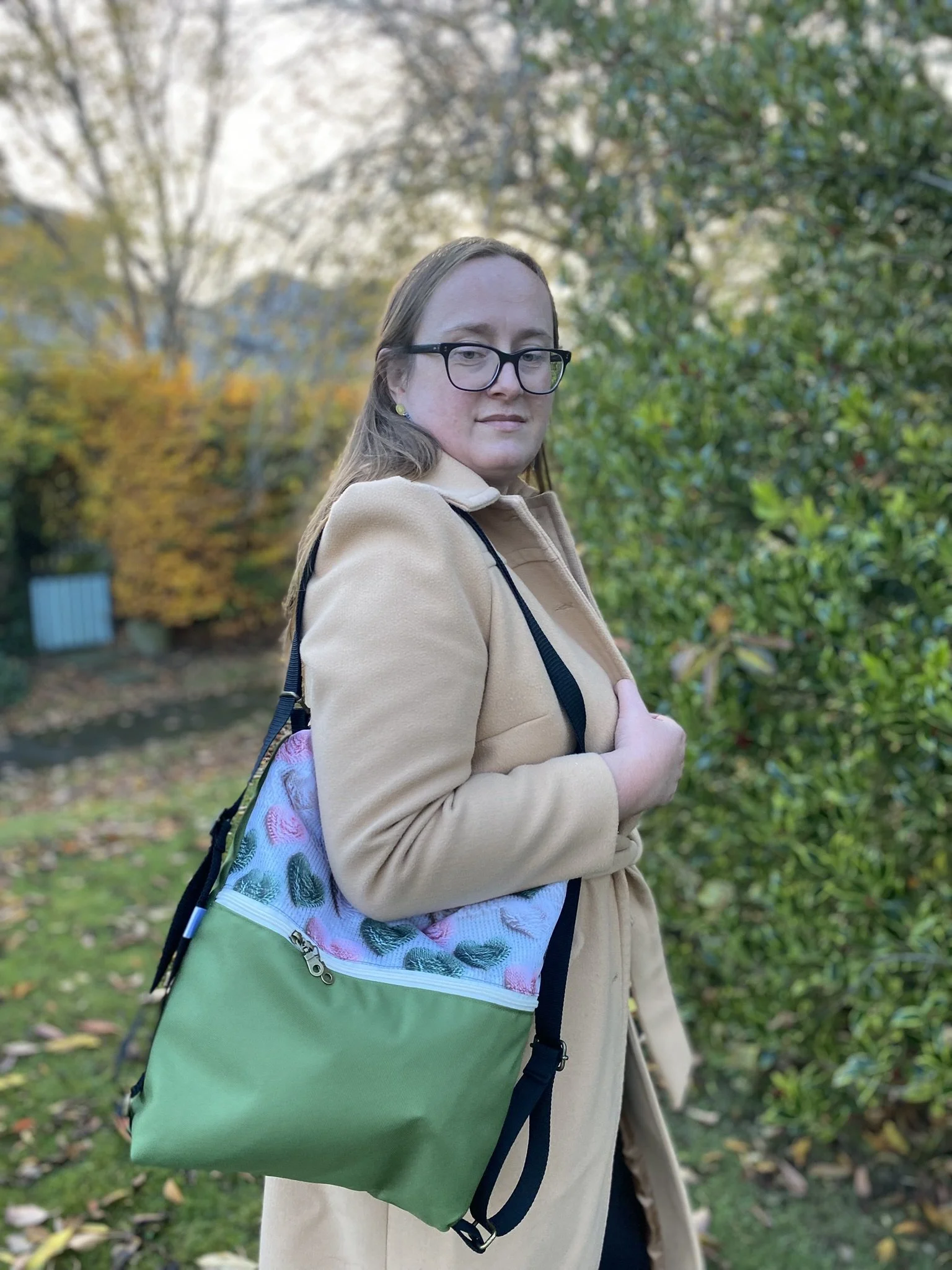 A woman with glasses and long light brown hair standing outdoors in a park during fall, wearing a beige coat and carrying a colorful, patterned tote bag over her shoulder.