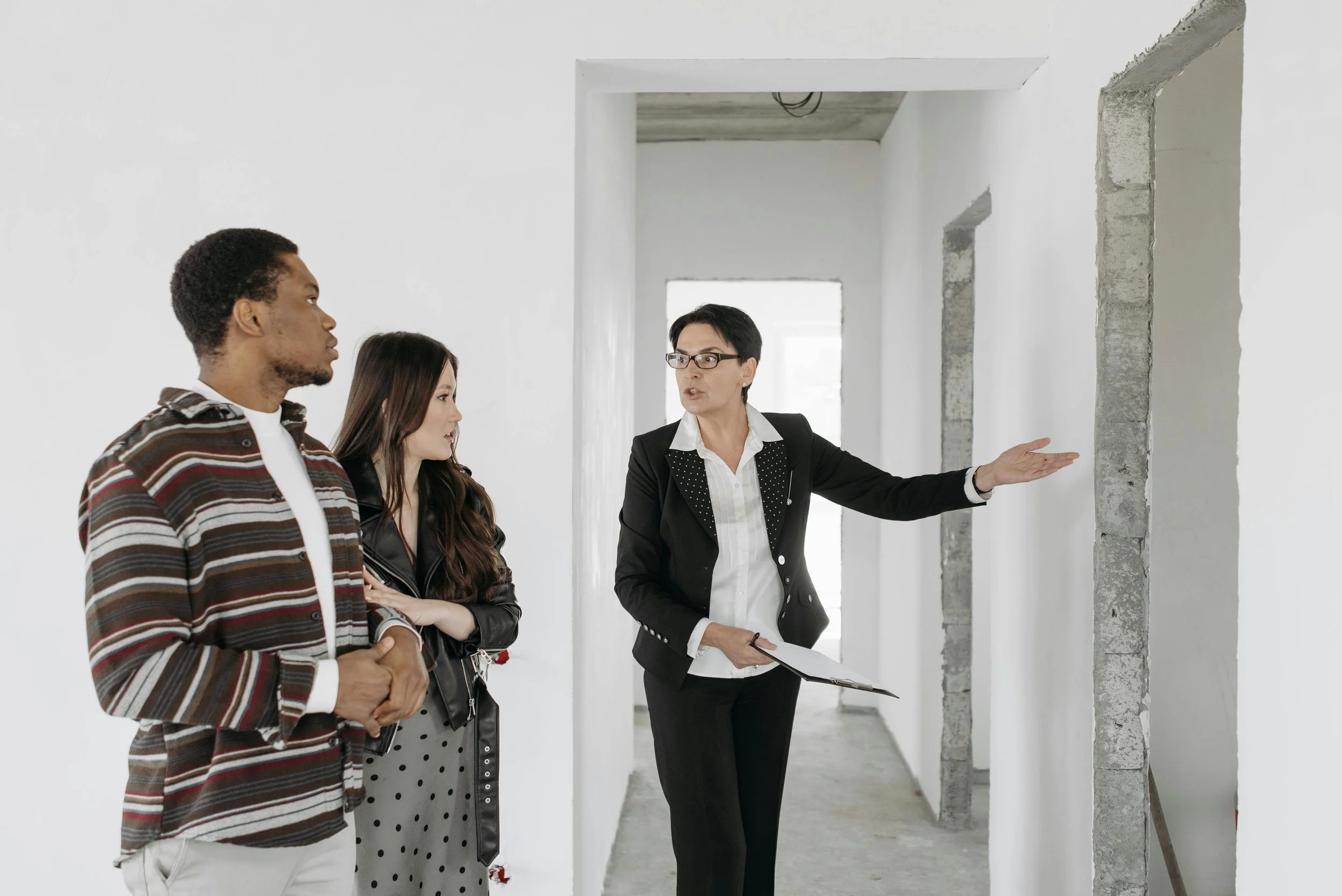 A woman in business attire explaining something to a young couple, standing in an unfinished building with white walls and exposed concrete frames.