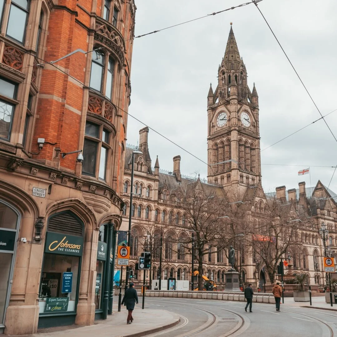 Street scene in front of historic stone building with clock tower, Gothic architecture, leafless trees, traffic lights, pedestrians walking, and storefronts, including Johnson's the Cleaners, with overhead tram wires and an English flag.