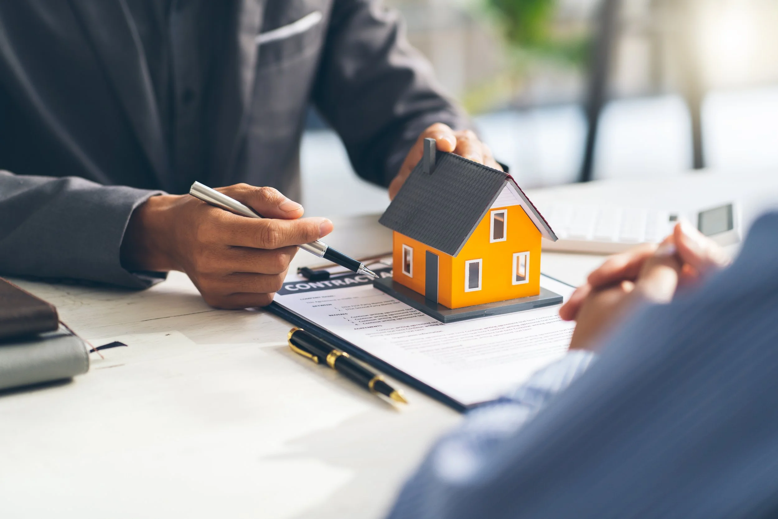 Two people sitting at a desk with a house model, contract, and pens, discussing home buying or real estate.