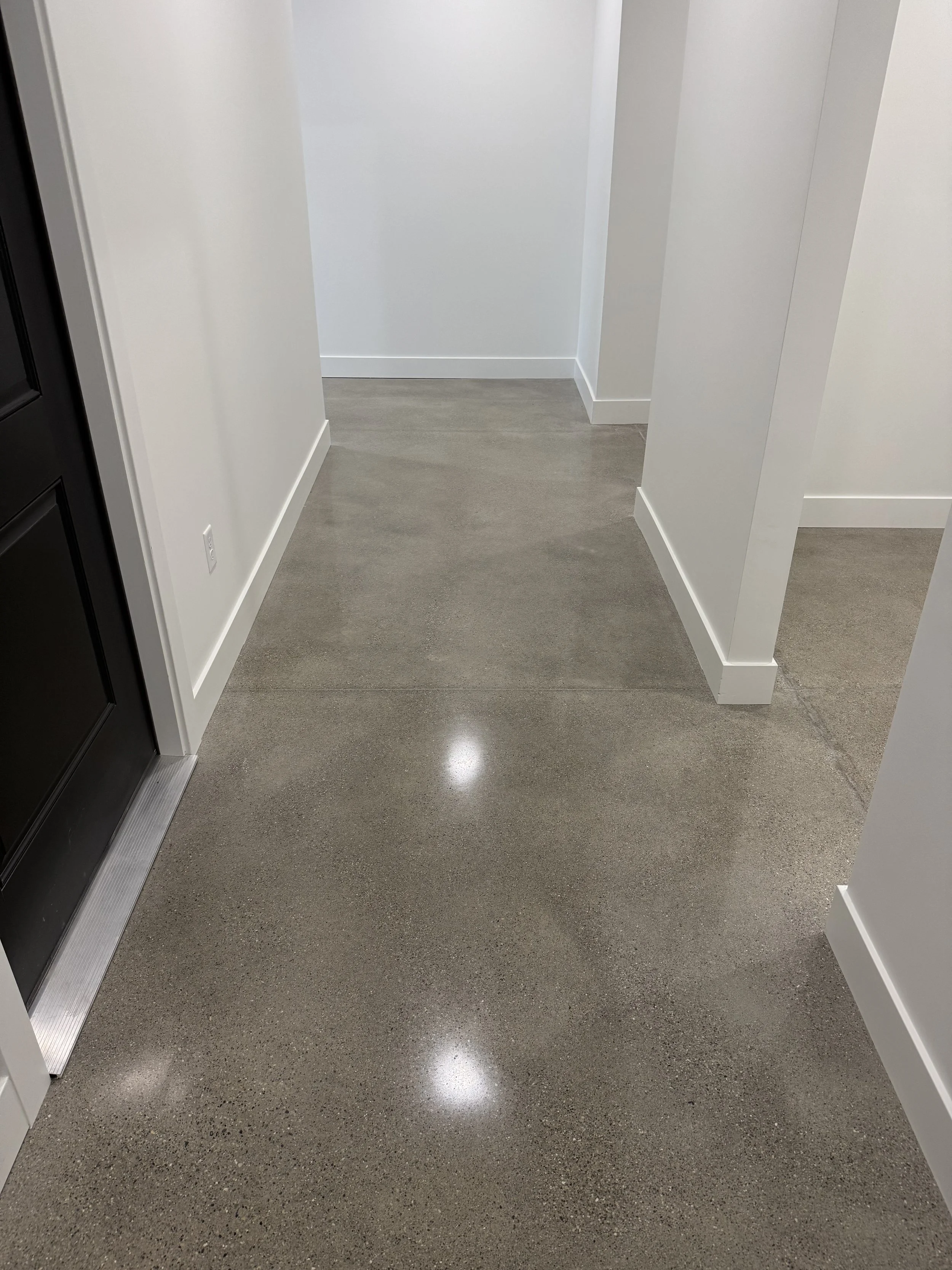 Polished concrete floor in a modern, minimal white-walled hallway with baseboards, near a black door.