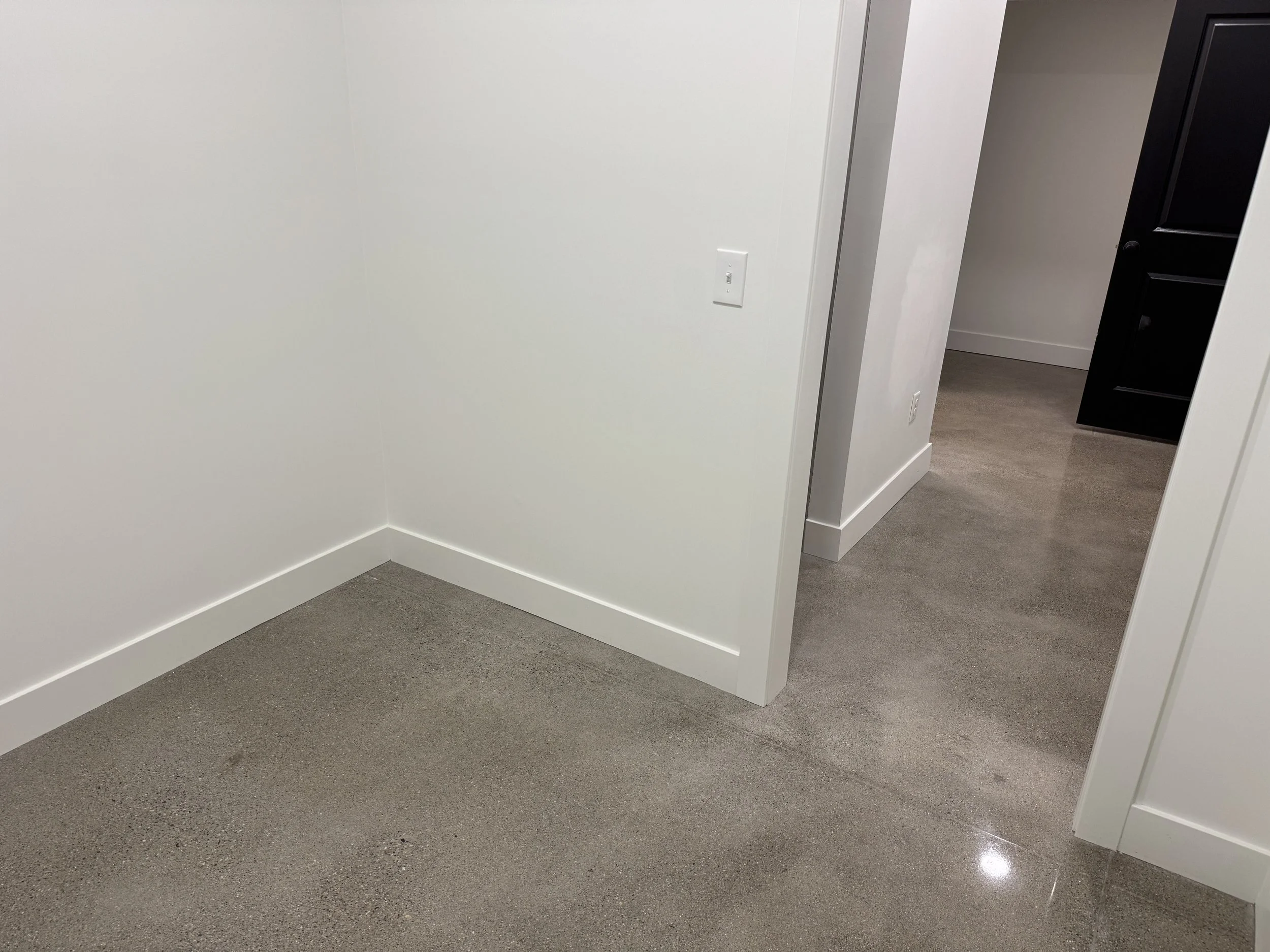Empty room with white walls, beige carpet, and a doorway leading to a hallway with dark flooring and black furniture.