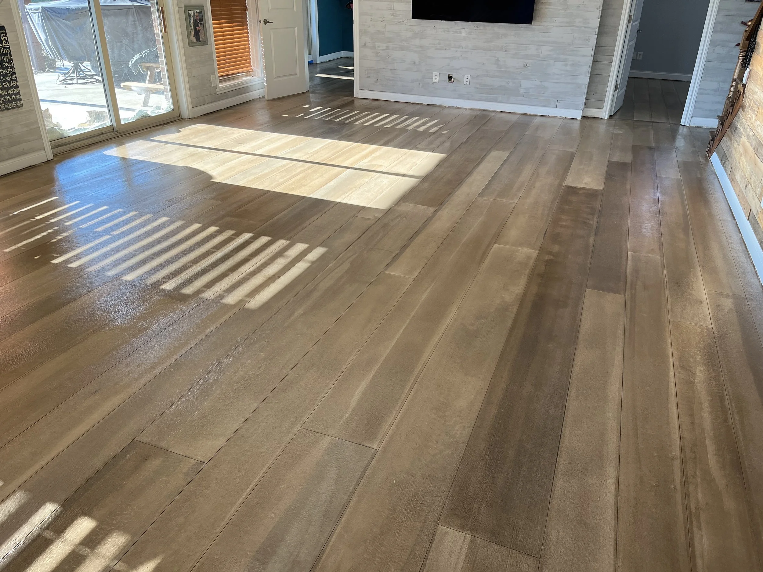 Living room with hardwood flooring, sunlight casting shadows through sliding glass doors, a white brick wall, and a television mounted on the wall.
