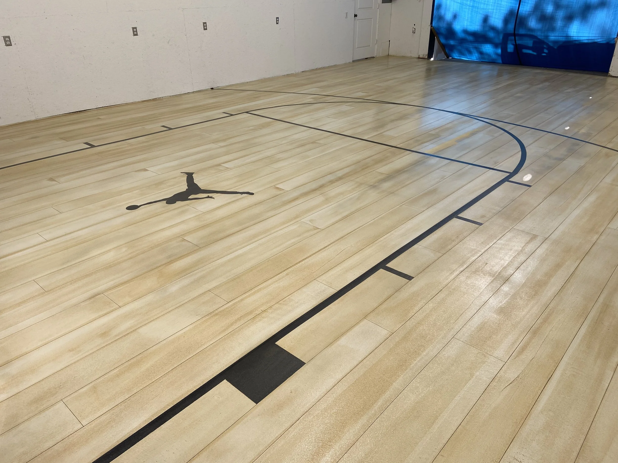 Empty indoor basketball court with a wood floor, a black silhouette of a person painted on the floor, and court lines in black and blue.