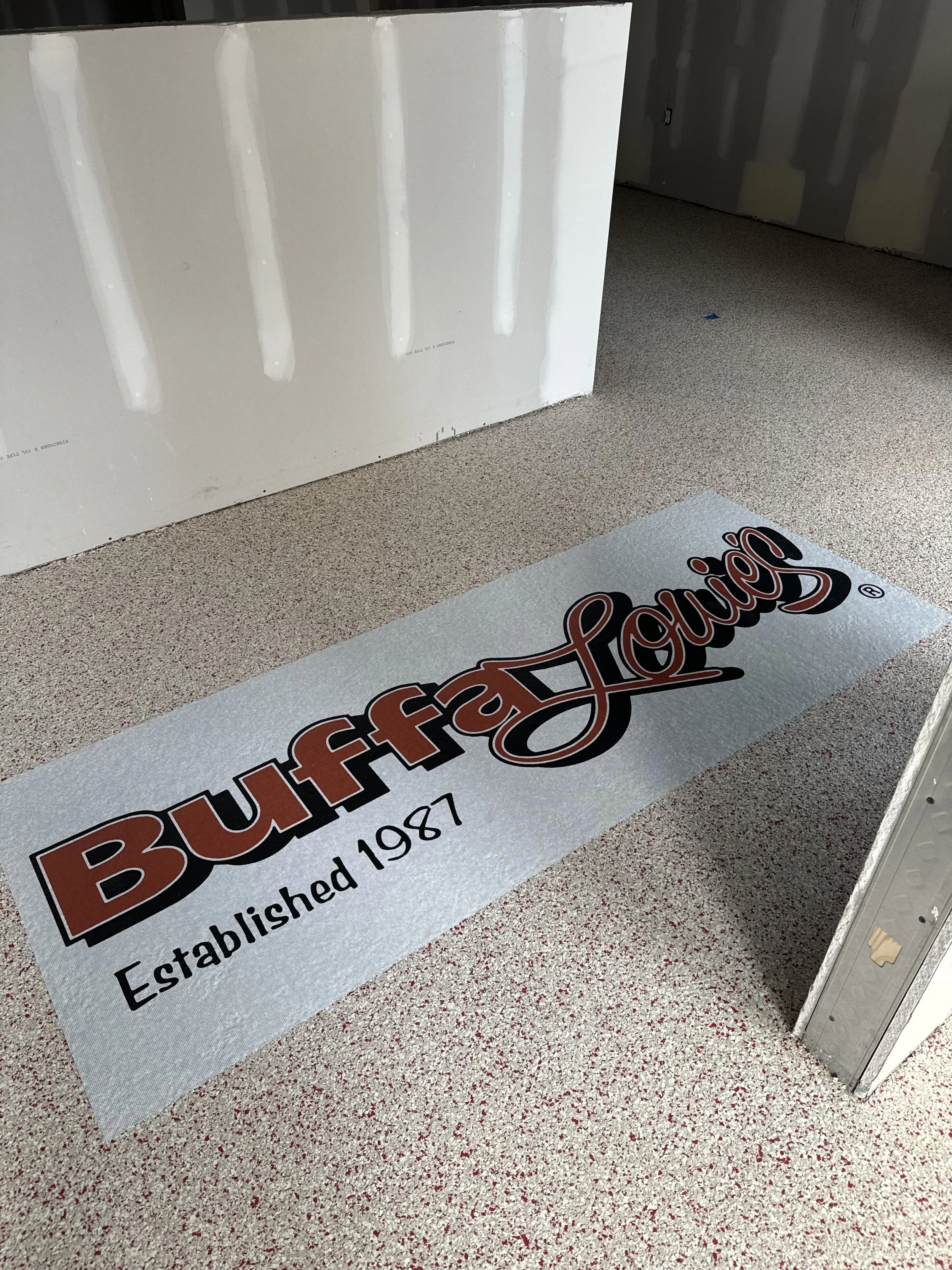 A printed floor mat with the BuffaLounge logo and the text 'Established 1987' on a speckled beige and red floor in a construction area.