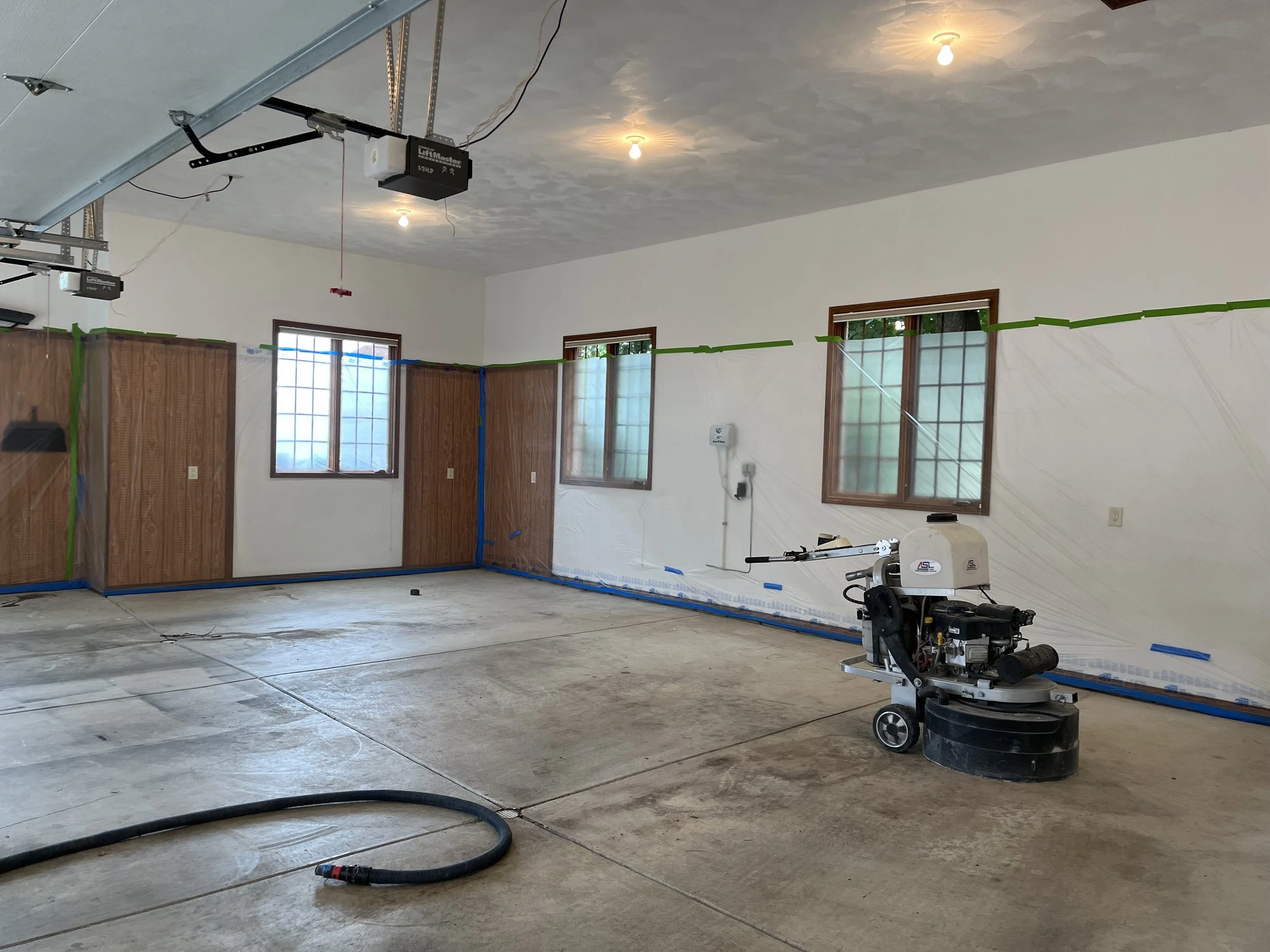 Garage or interior room with concrete floor, construction or renovation in progress, with protective sheeting on walls, windows with wooden trim, ceiling with exposed light bulbs, large flooring polishing machine, and garage door mechanisms visible.