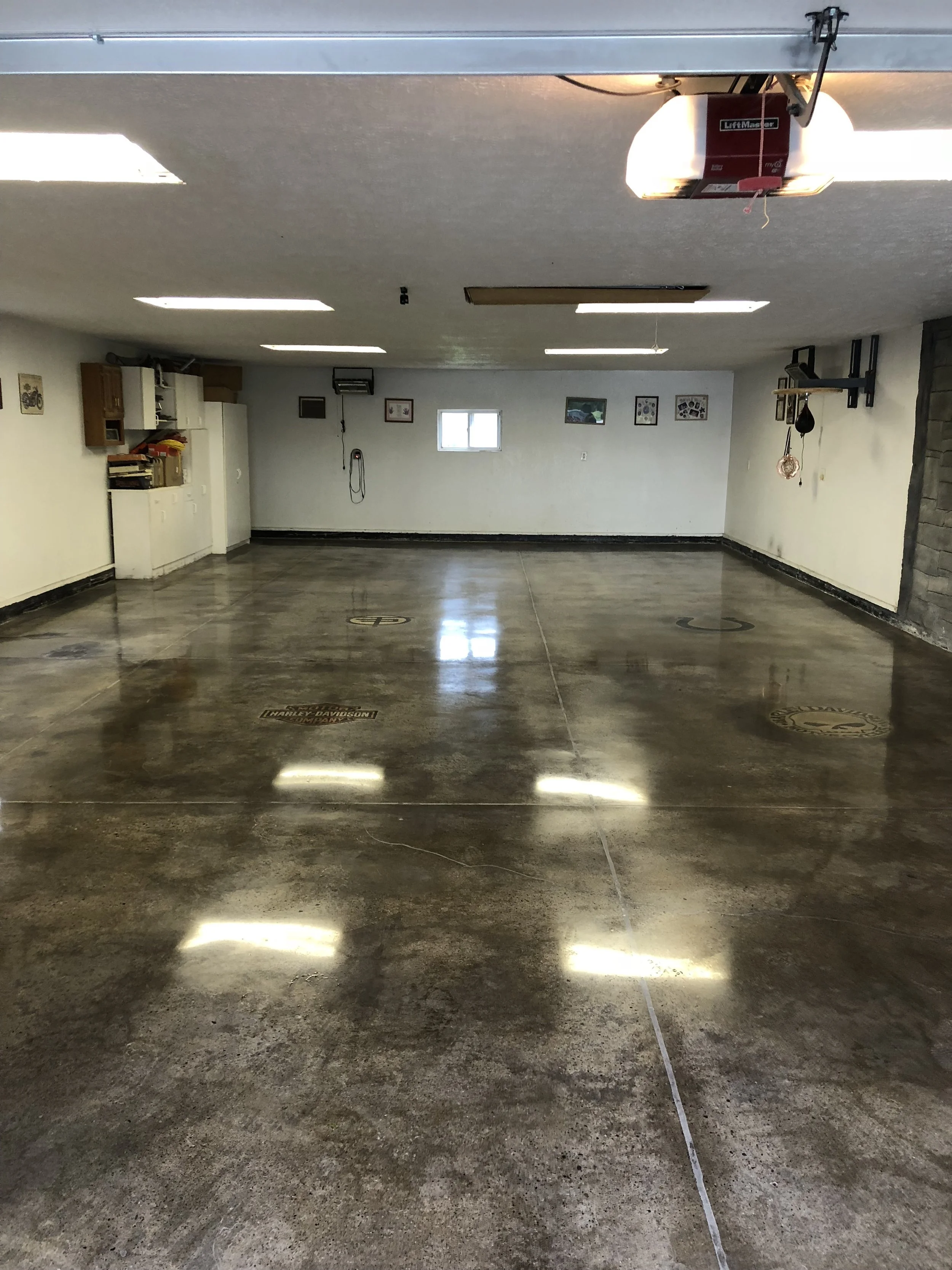 Empty garage with a polished concrete floor, garage door opener on the ceiling, small windows on the back wall, and wall-mounted shelving and tools.