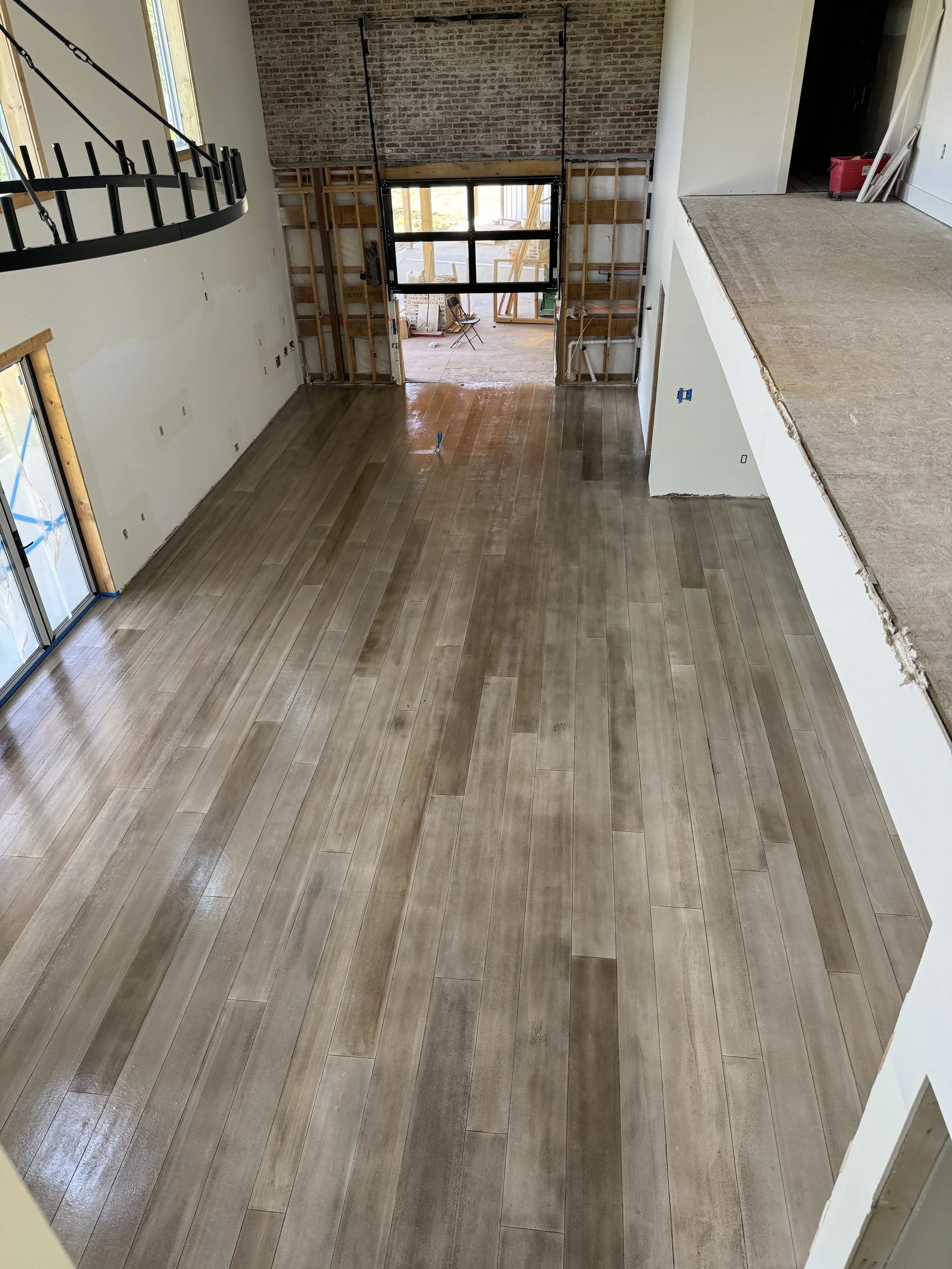 Empty living room under construction with new wood flooring, large window, exposed brick wall, and a partially built wall near the entrance.
