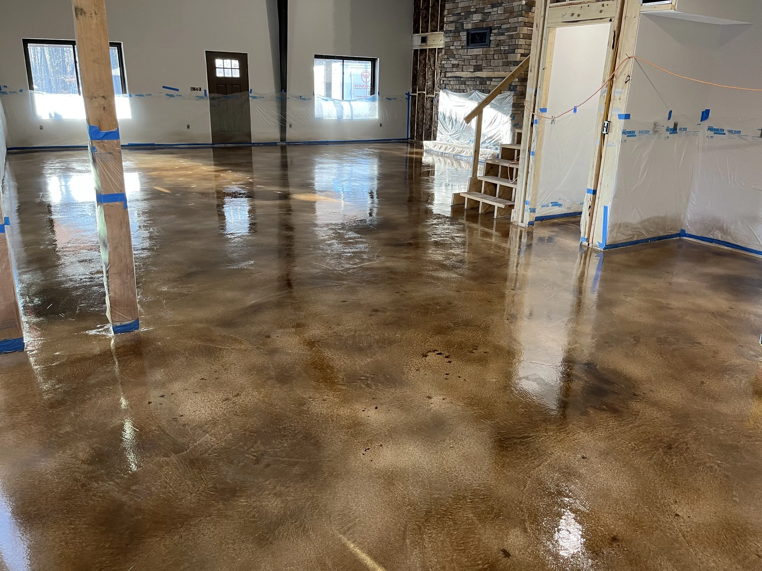 Interior of a house under construction with a freshly stained polished concrete floor, partial wall framing, plastic coverings, and windows.