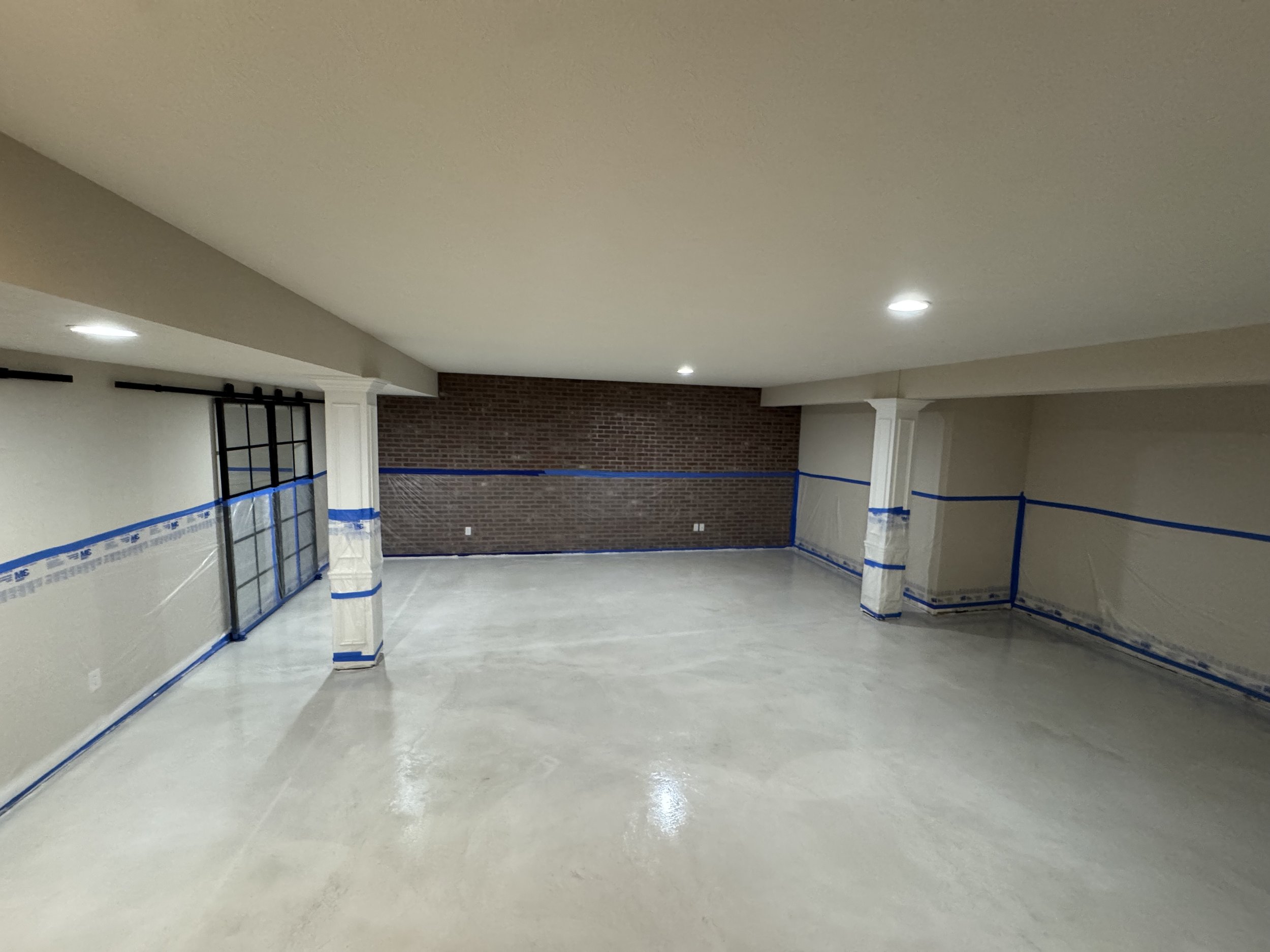 Empty room with white walls, brick accent wall, ceiling lights, and taped plastic covering the lower walls.