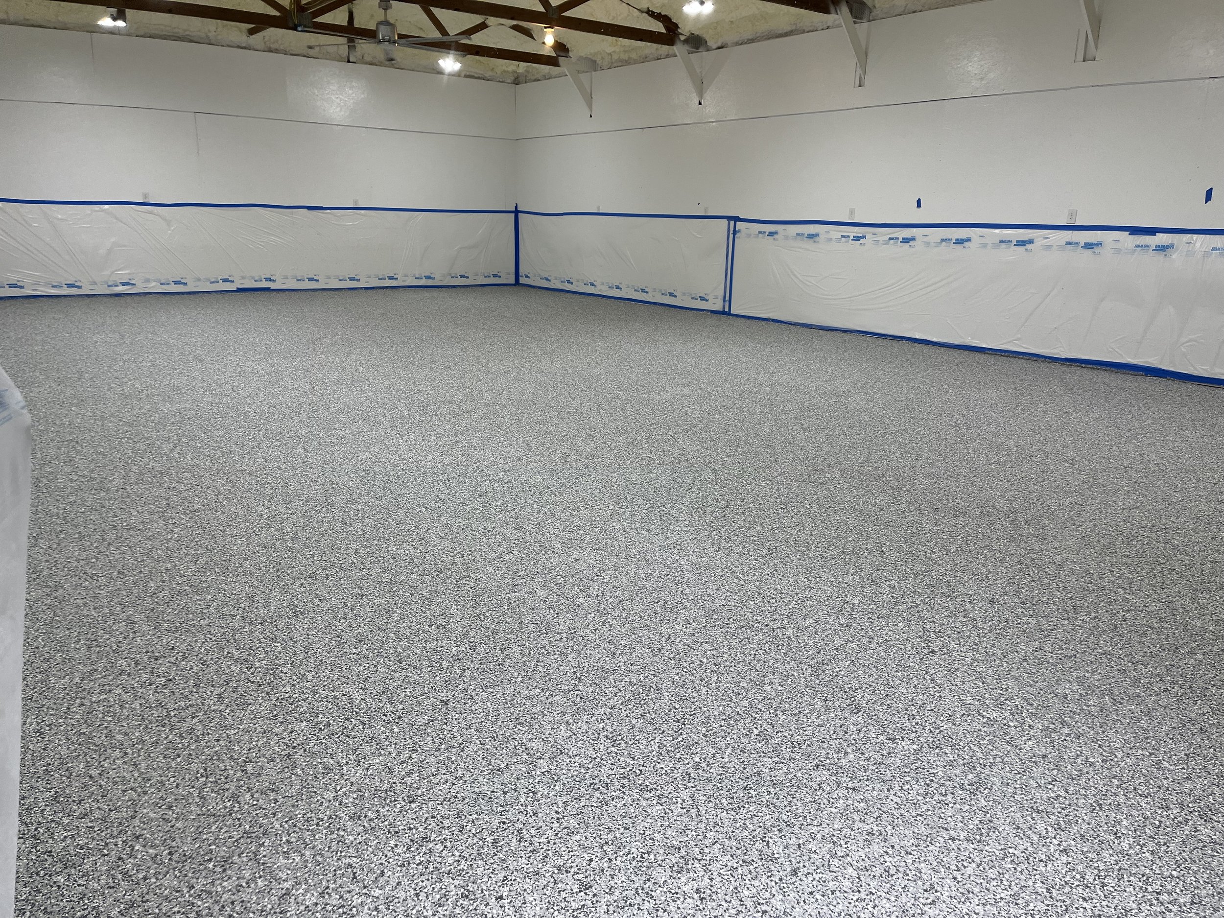 Empty indoor room with unfinished ceiling, gray speckled flooring, white walls covered with plastic sheeting, and blue tape lining the walls.