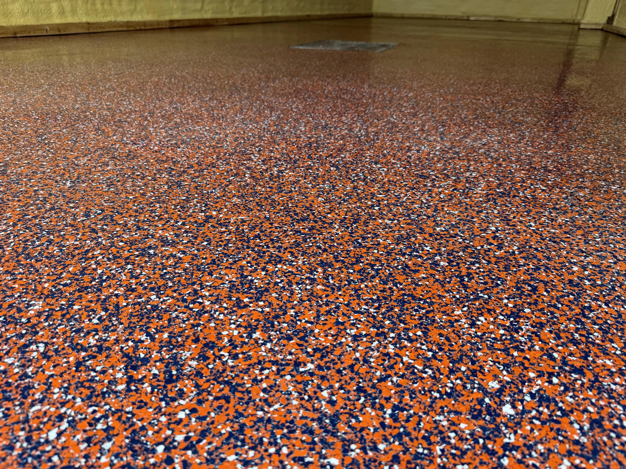 Close-up of a speckled orange, black, white, and blue rubber gym floor with a floor drain in the background.