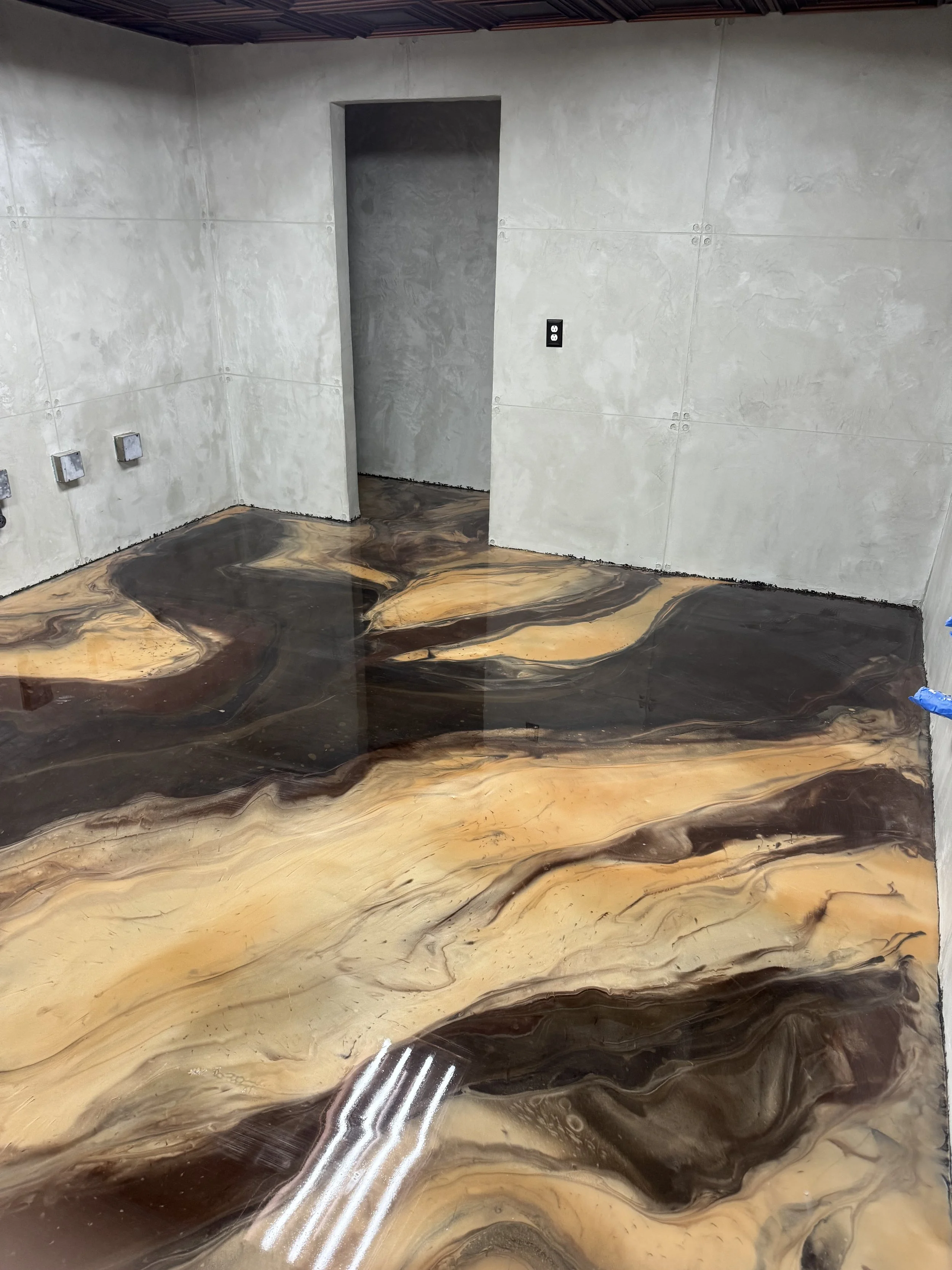 Room with an epoxy resin floor featuring swirls of black, brown, gold, and beige colors. The walls are unfinished with visible seams and a doorway on the back wall. There are electrical outlets on the left wall and a black electrical outlet on the back wall.
