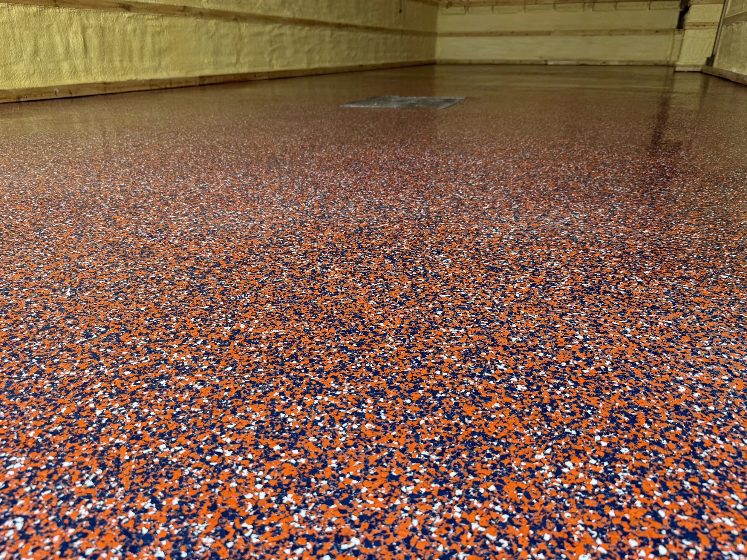 Close-up view of a speckled orange, blue, black, and white epoxy-coated floor near a yellow wall and floor drain.