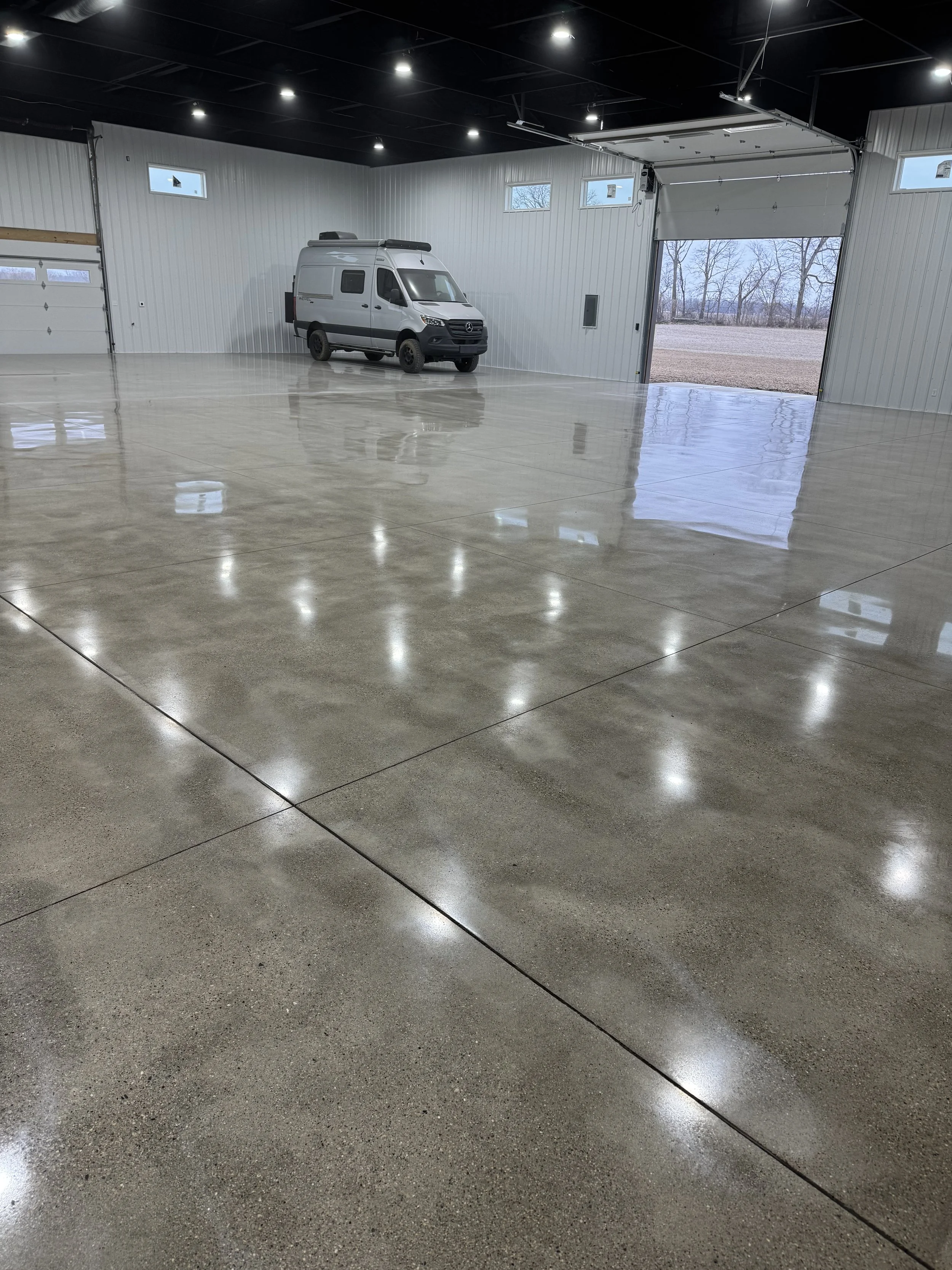 Empty, polished concrete floor inside a large garage with a white van parked near the wall and open garage door showing an outdoor view of trees and grass.