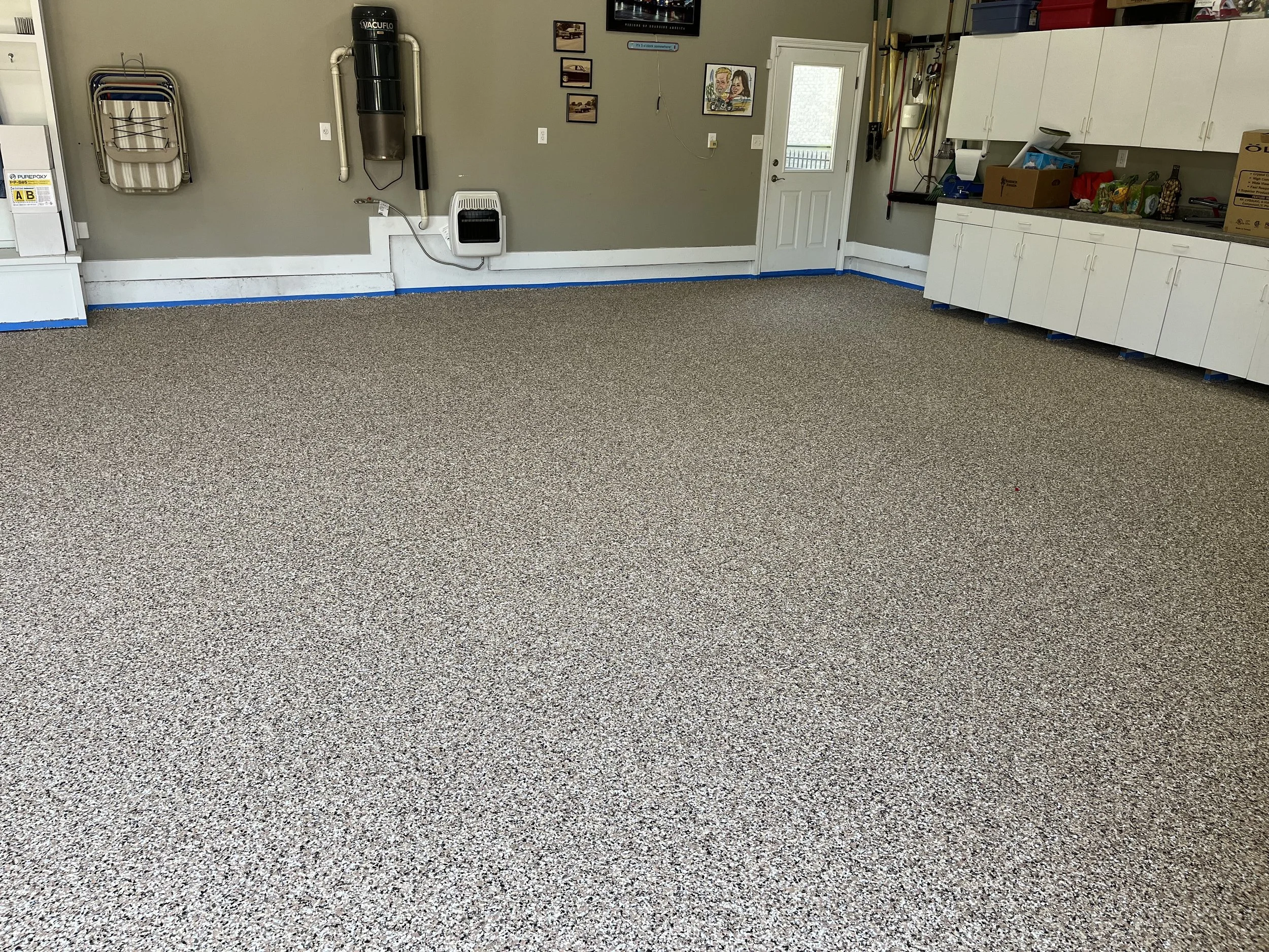 Empty garage or workshop space with speckled beige carpet, white cabinets on the right, a door at the back, and various tools and equipment on the wall.