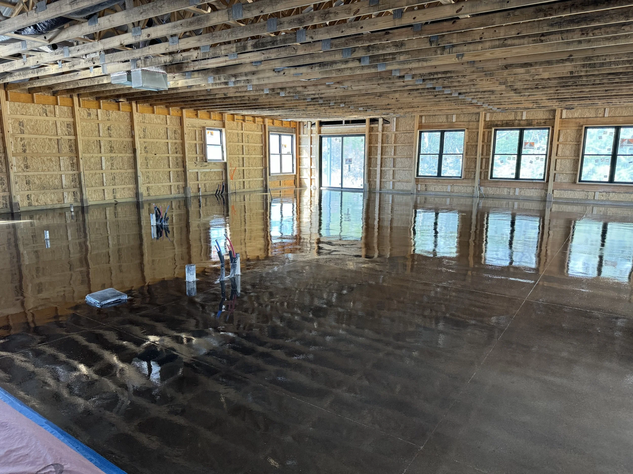 Interior of a building under construction with a polished concrete floor and exposed wooden framing, multiple windows, and ceiling joists visible.