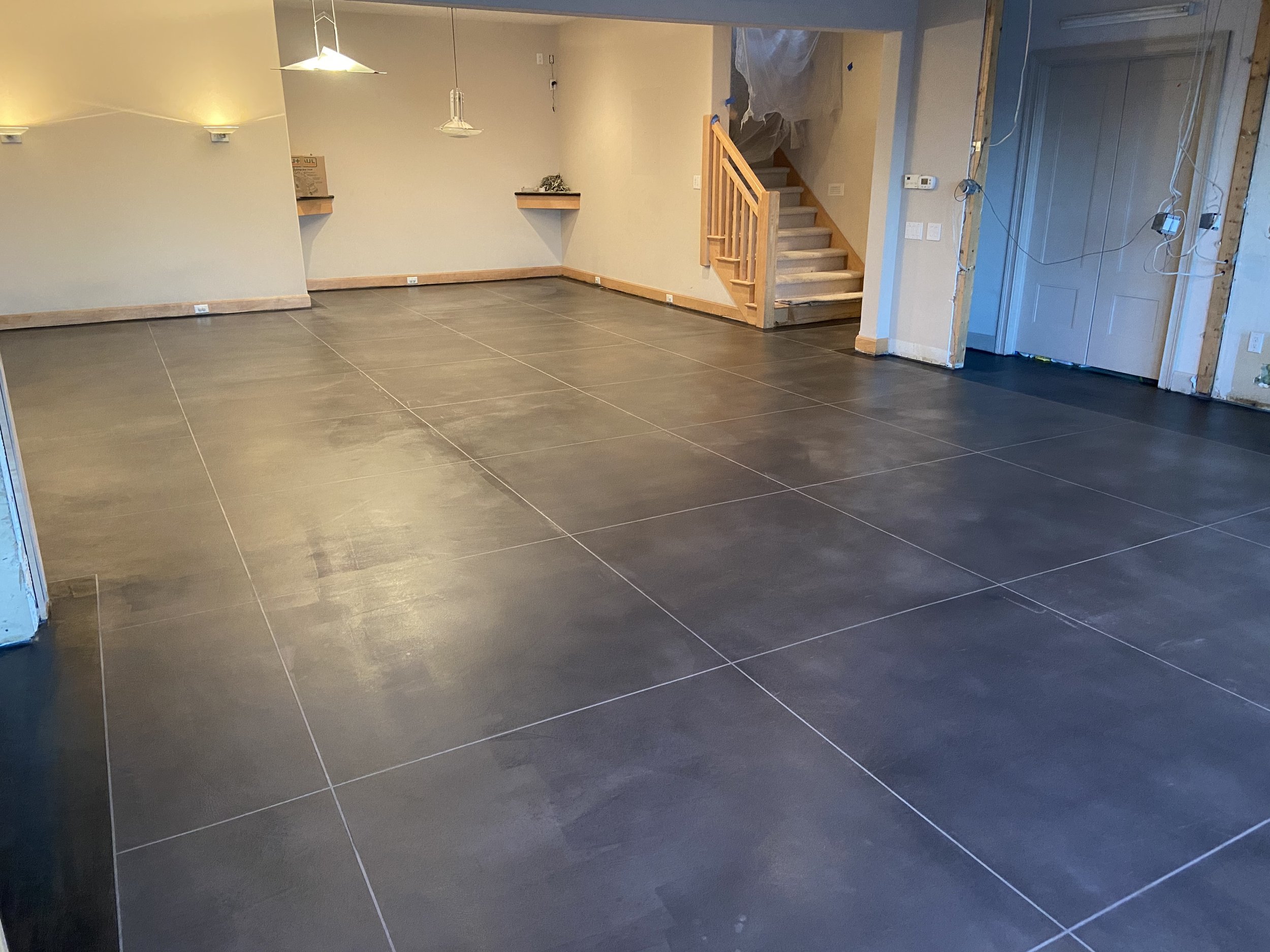 Empty room with new dark tiled flooring, beige walls, and wooden trim. Construction or renovation in progress with exposed wiring and unfinished wall sections. Staircase with wooden railing leads to an upper level.
