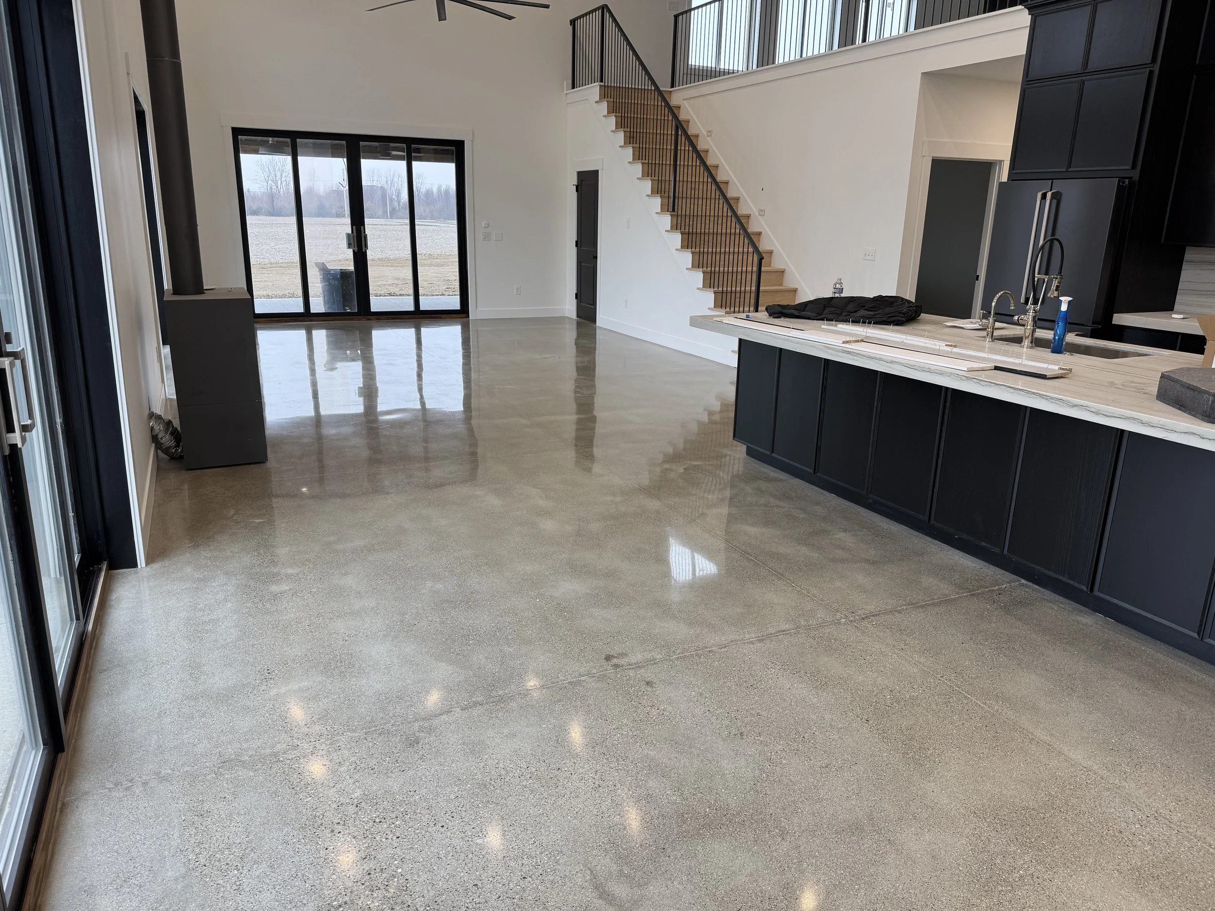 Empty modern kitchen and living area with polished concrete floors, black cabinets, and a staircase with wooden steps and black railing.