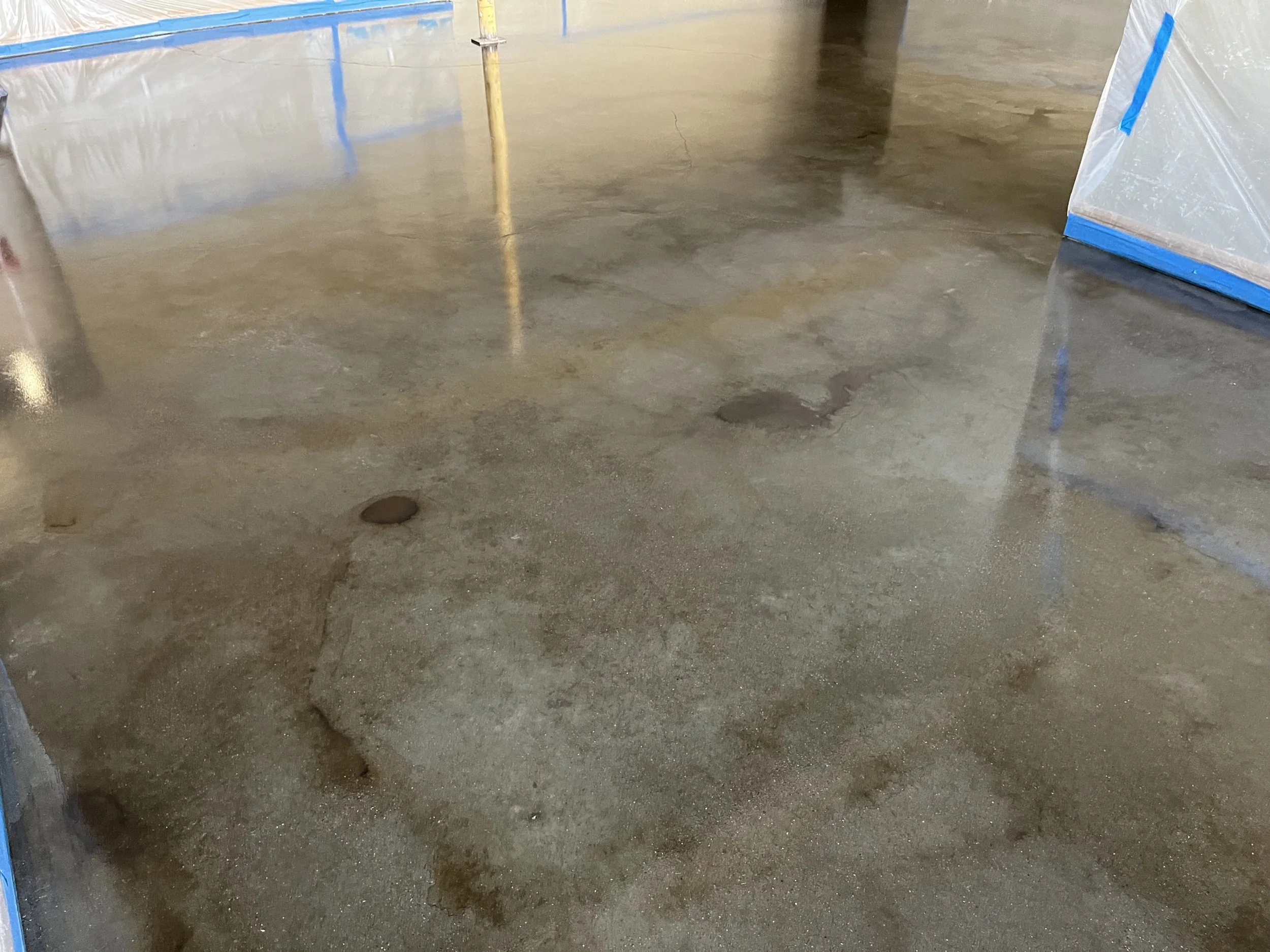 Concrete floor with wet patches and floors drains, surrounded by plastic barriers with blue tape.