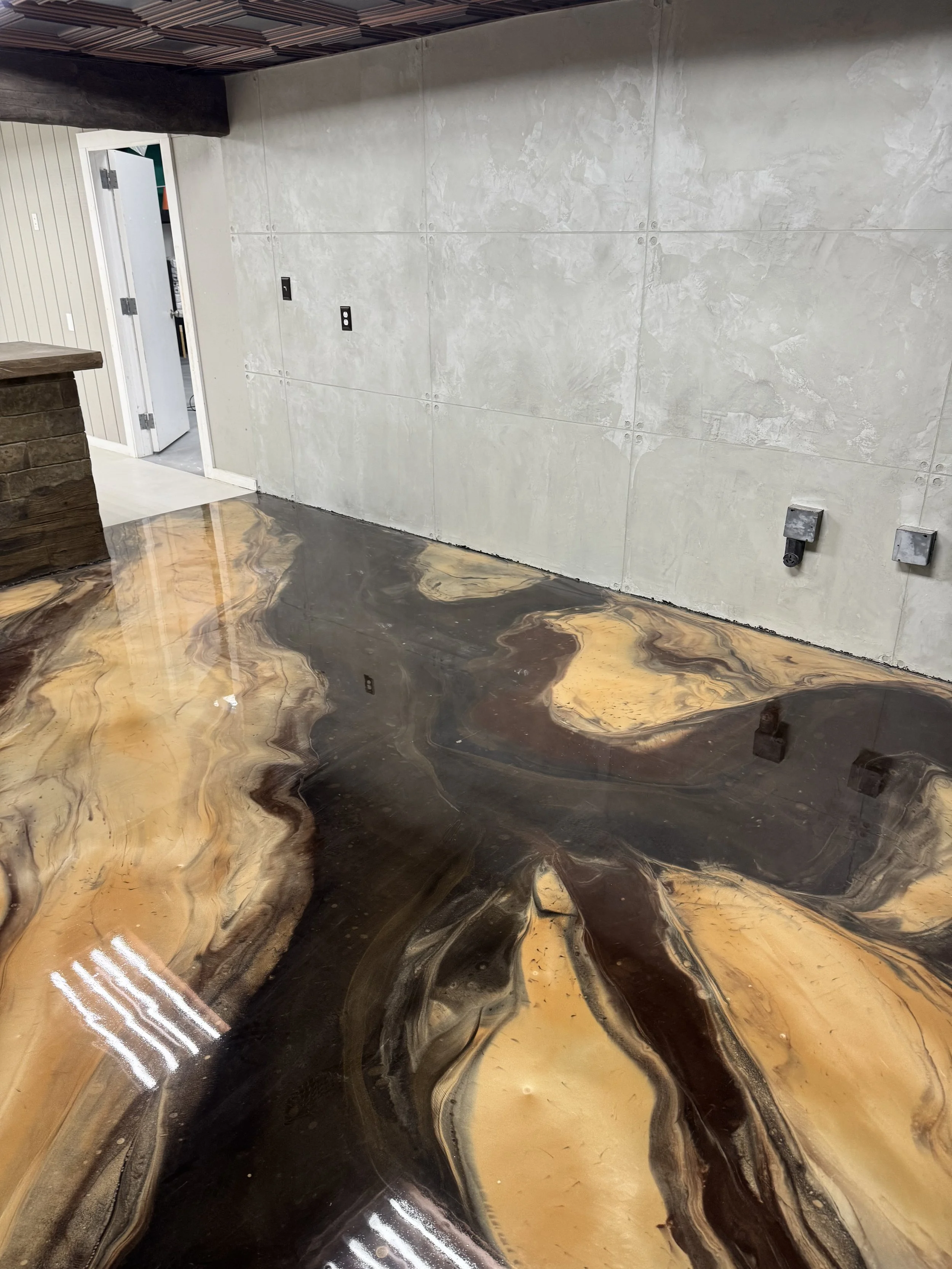 Interior room with abstract-patterned polished concrete floor in tan, brown, and black colors, plain light-colored walls, some electrical outlets and switches, and a door open to another room.