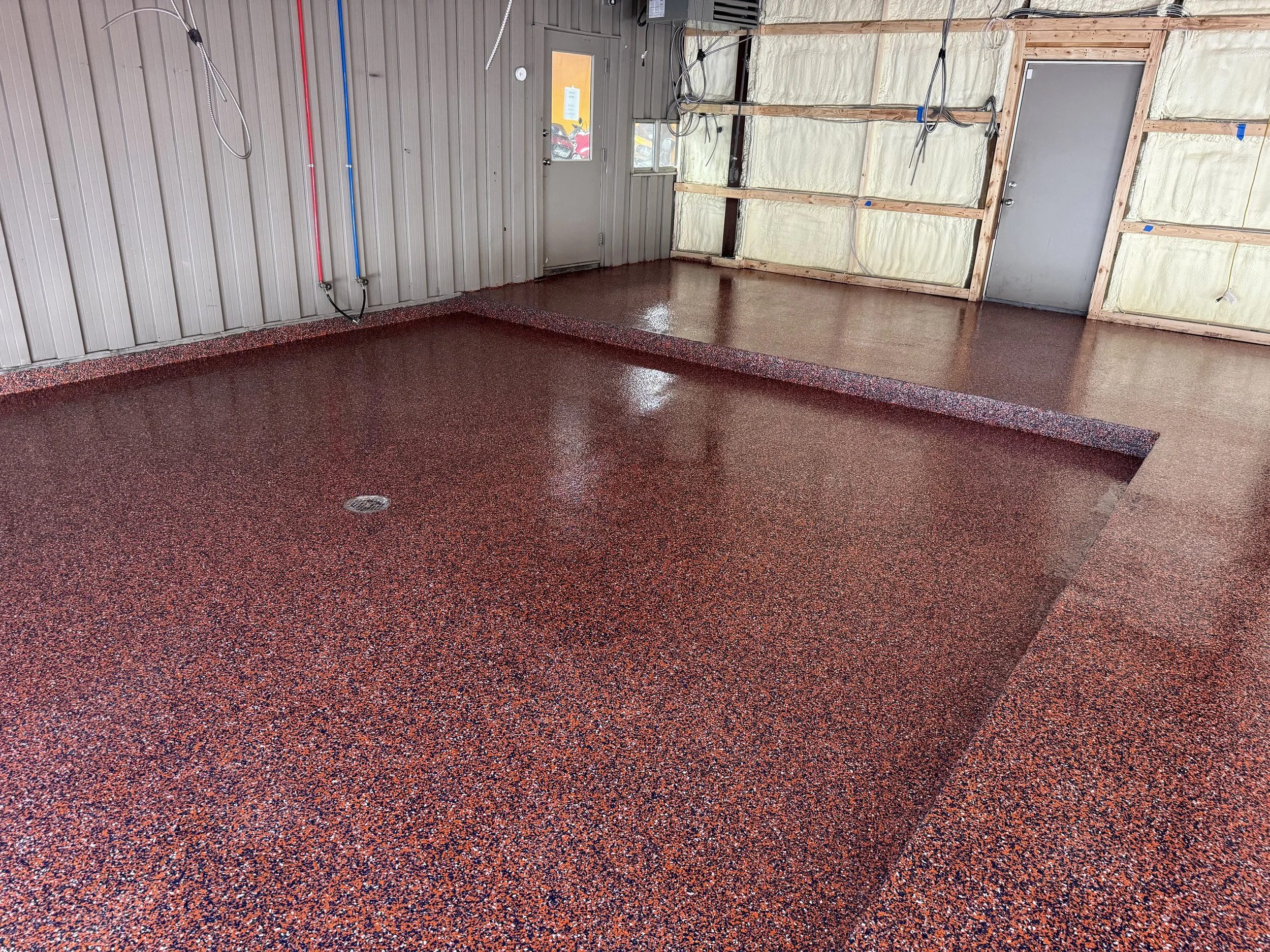 An indoor space under construction with a red and black speckled epoxy floor, exposed wall framing, and an unfinished corner with insulation and wiring.