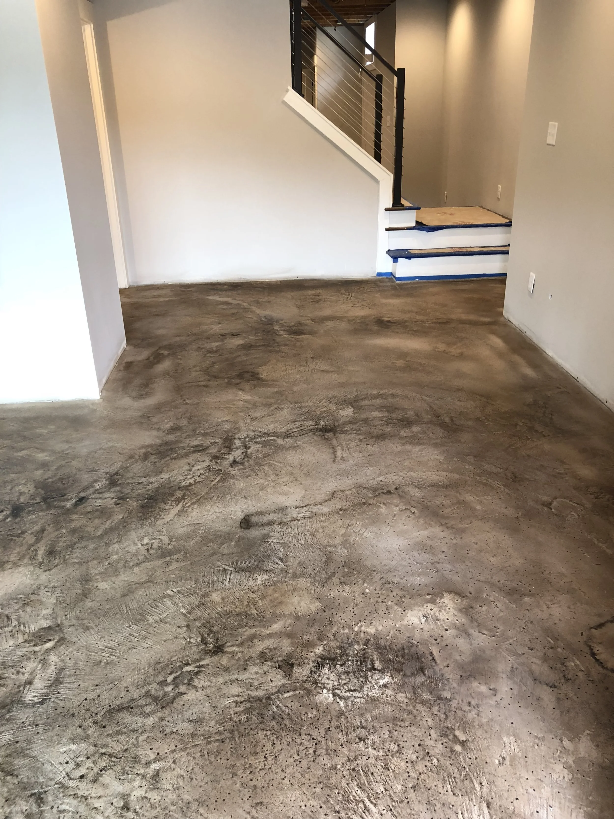 Interior of a house with unfinished concrete floor, staircase with wooden steps and black metal railing, and light-colored walls.