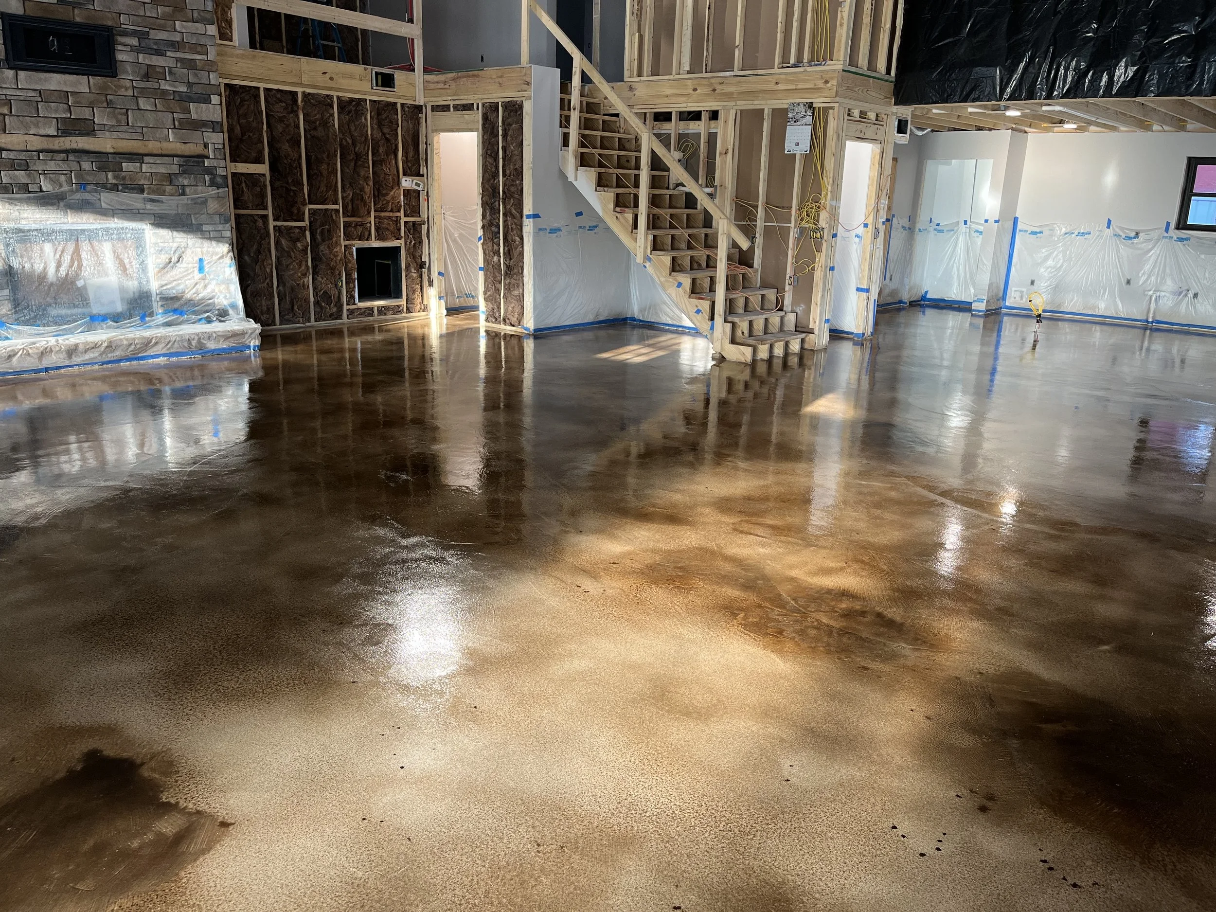 Interior view of a newly constructed home with an unfinished staircase, open floor with a freshly poured epoxy coating, and plastic sheeting protecting parts of the walls.