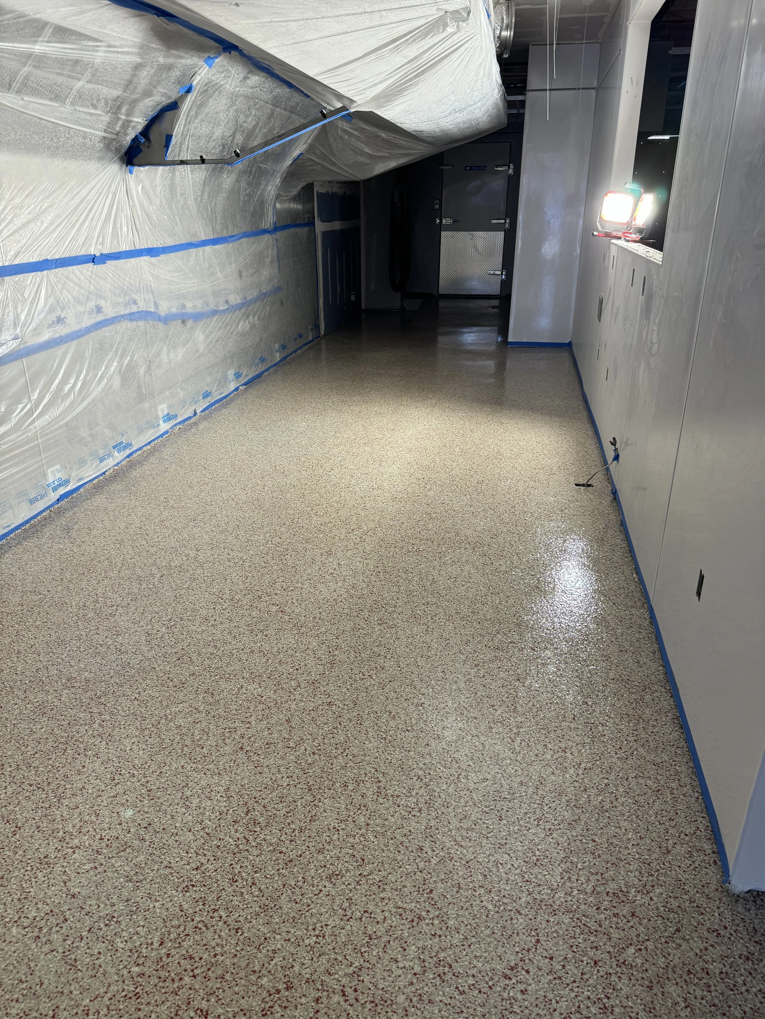 Empty hallway under construction with plastic sheeting on the ceiling, partially painted walls, and a shiny terrazzo floor.
