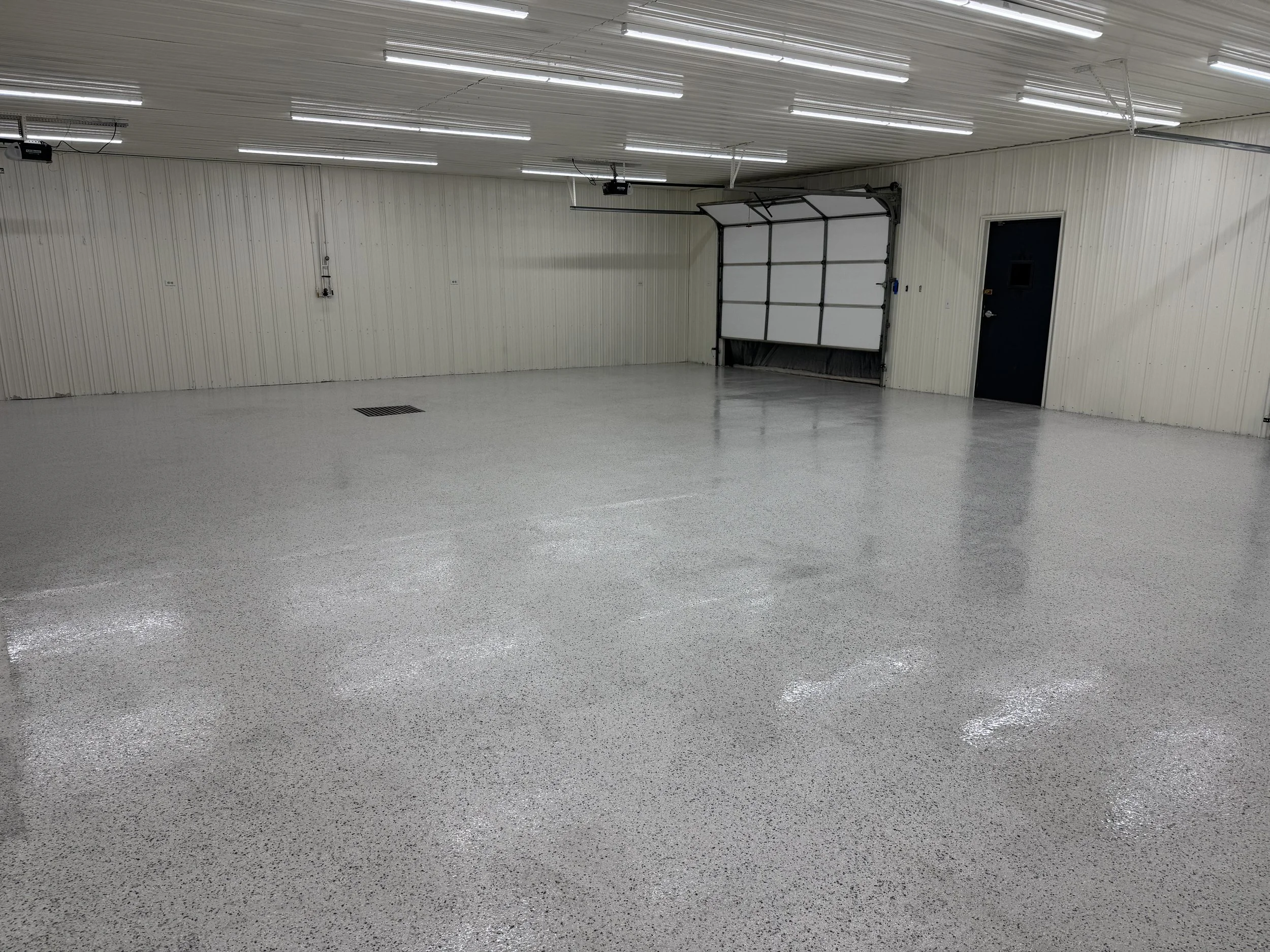 Empty, clean garage with white speckled epoxy floor, beige paneled walls, and ceiling with fluorescent lighting. Garage door on the far wall, black door on the right wall, and wall-mounted electrical panel.