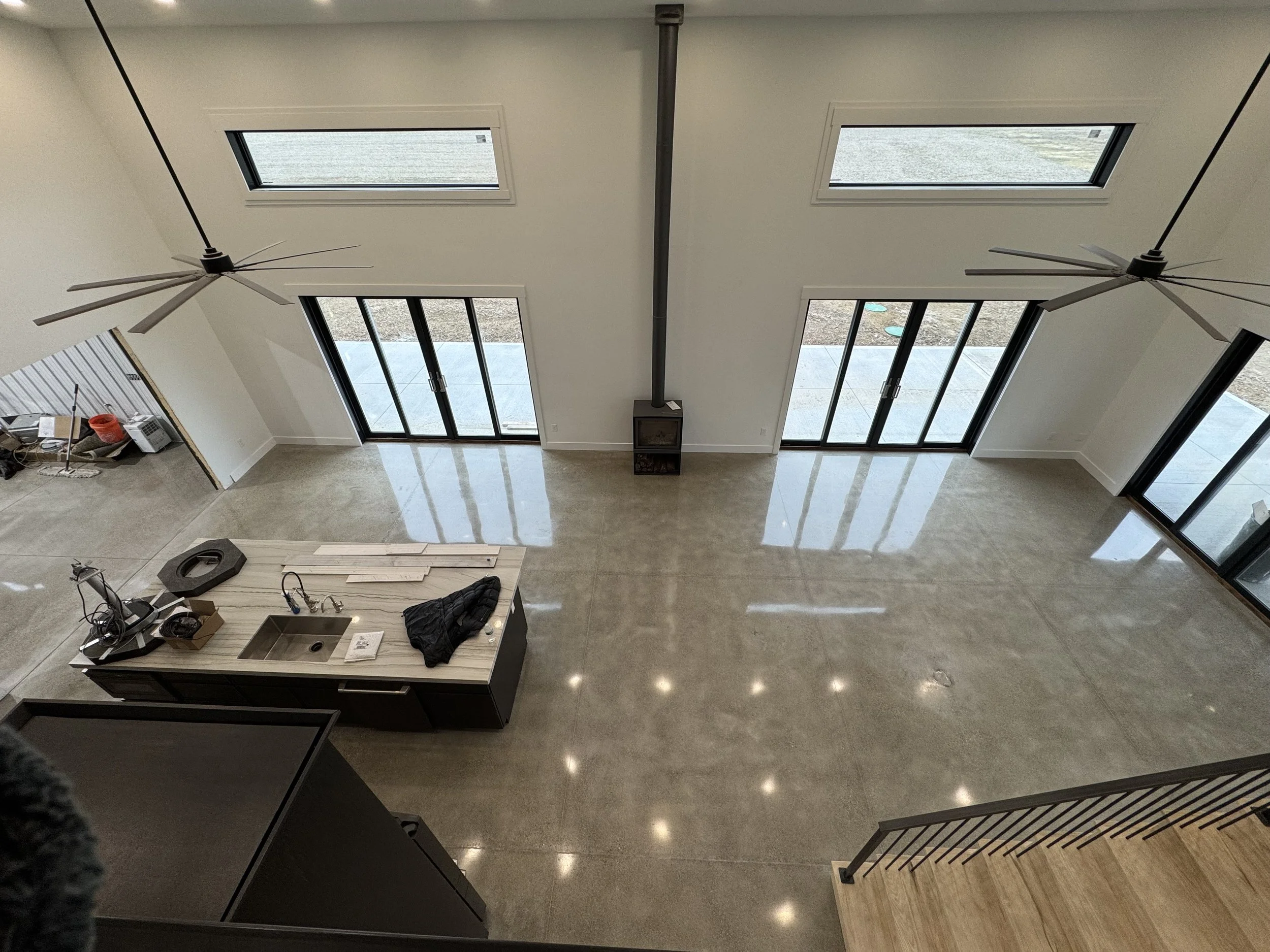 An empty modern living room with large glass sliding doors and windows, two ceiling fans, and a kitchen island with various items on it. The room has a polished concrete floor and high ceiling.