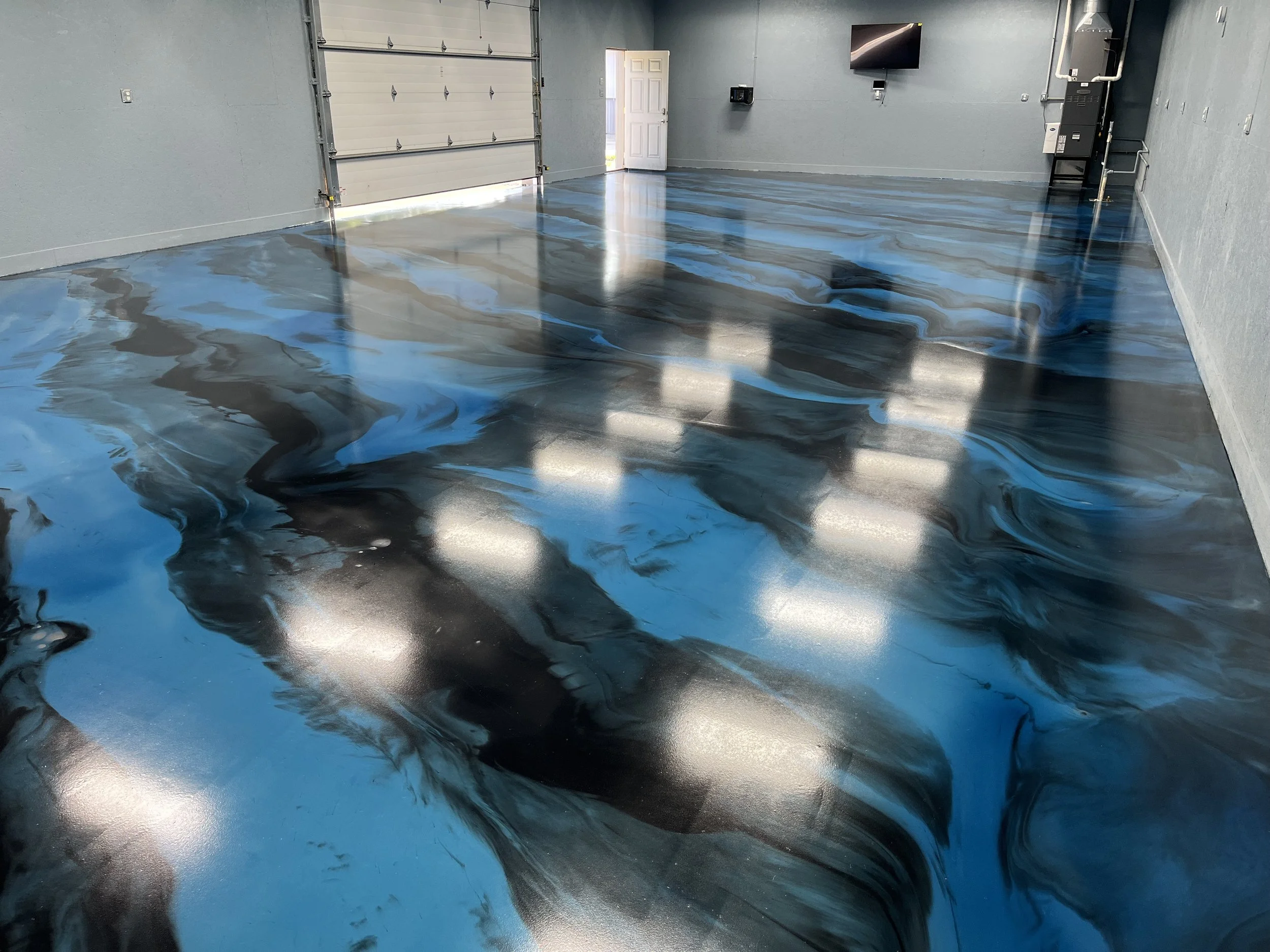 Empty garage with a glossy, abstract-patterned epoxy floor in shades of blue, black, and gray, with an open garage door and a wall-mounted TV.