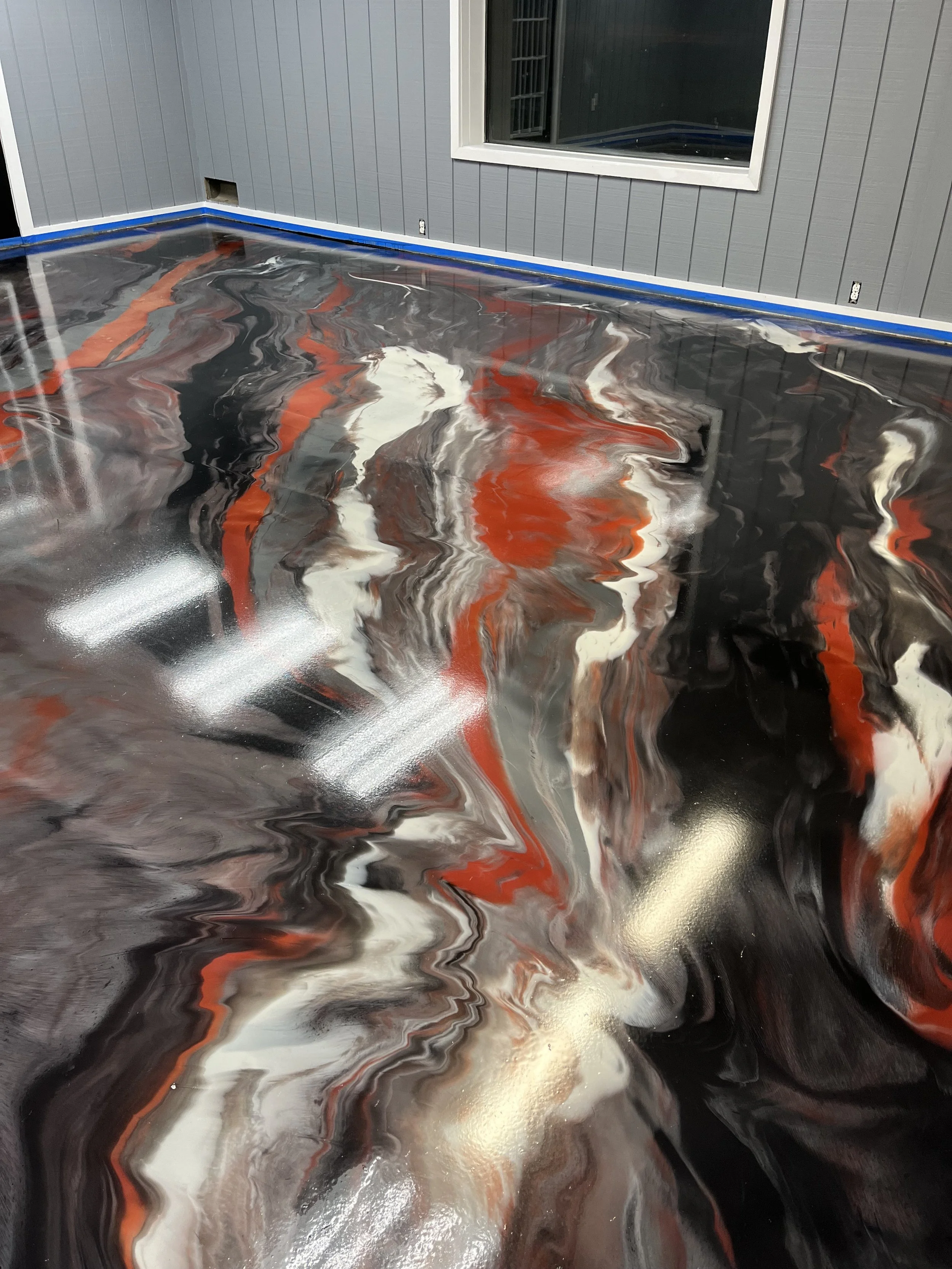 Close-up of a polished epoxy floor with swirling patterns in black, white, gray, and red, located inside a room with blue trim and gray wall paneling.