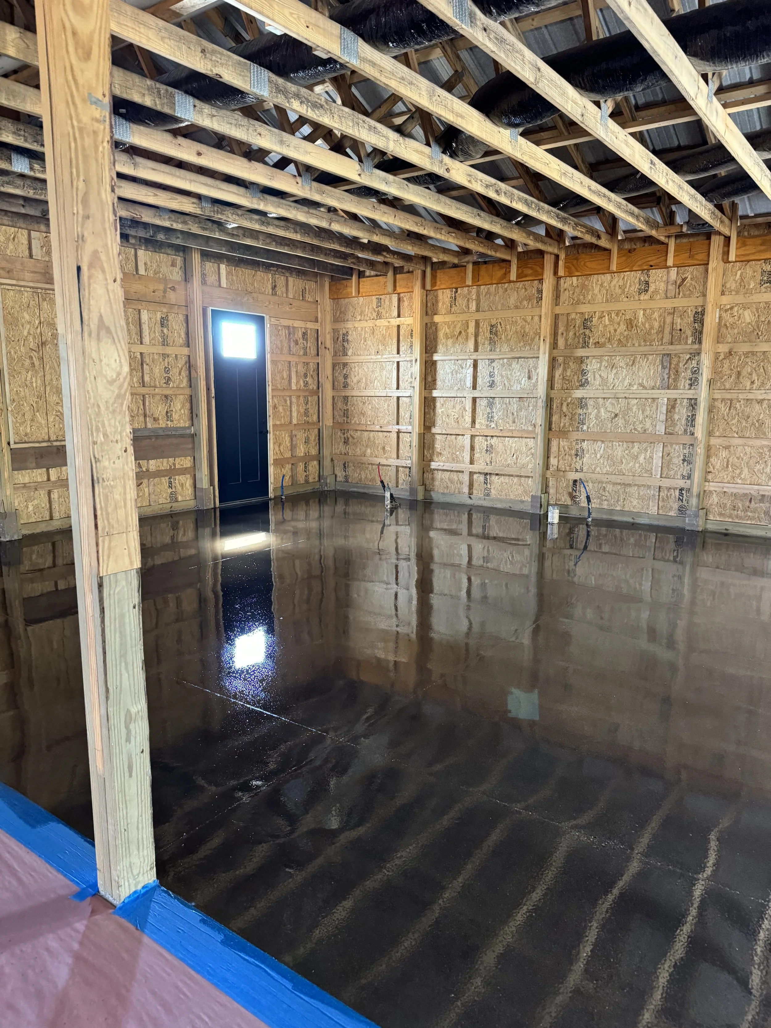 Interior of a house under construction with a concrete floor that has just been finished, showing fresh wet concrete. The walls are framed with wood and the ceiling shows exposed wooden beams and insulation. A black door with a small window is visibl