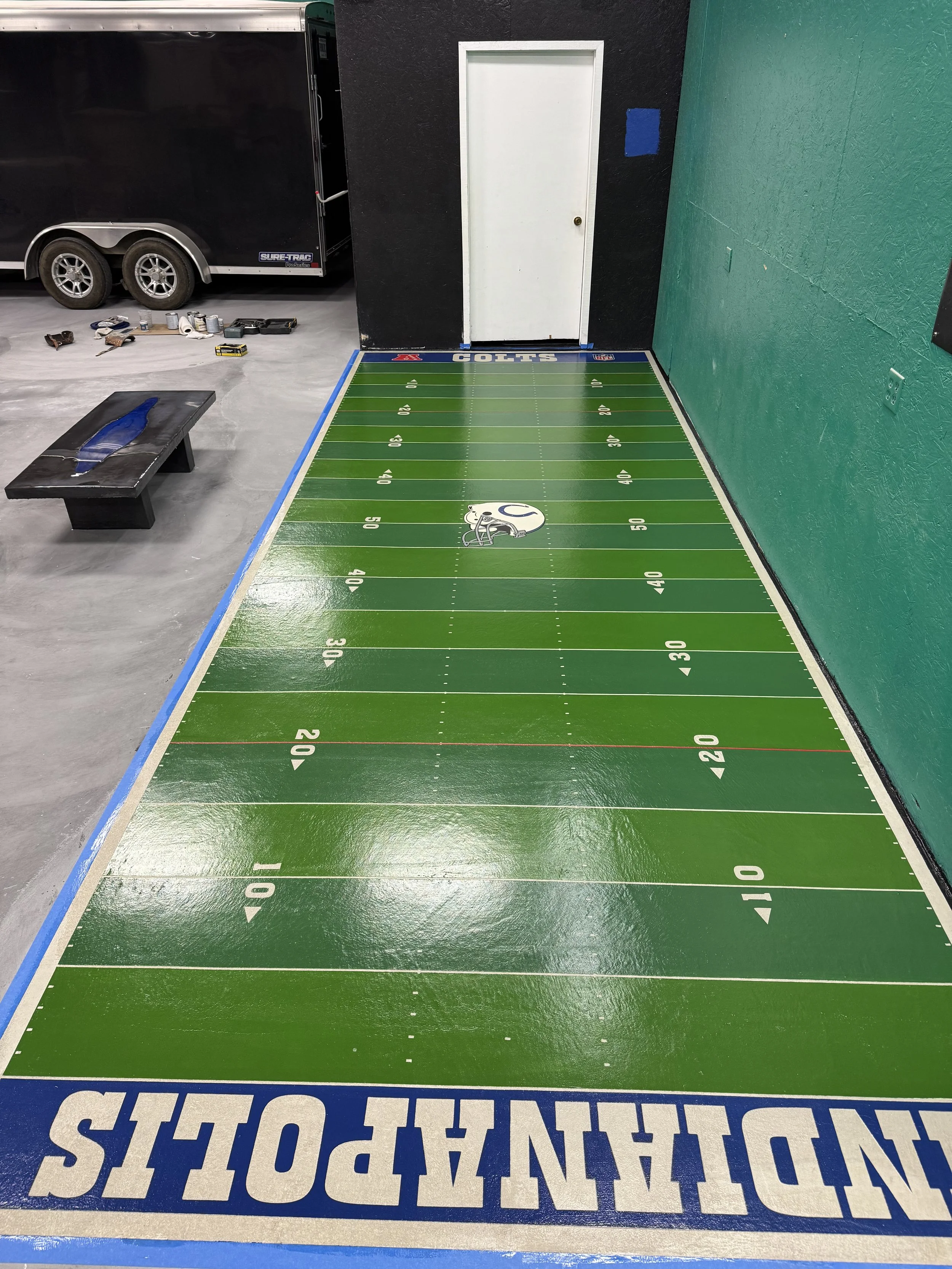 Indoor mini football field with the Indianapolis Colts logo at the center and team name along the end zone, situated against a green wall with a white door.
