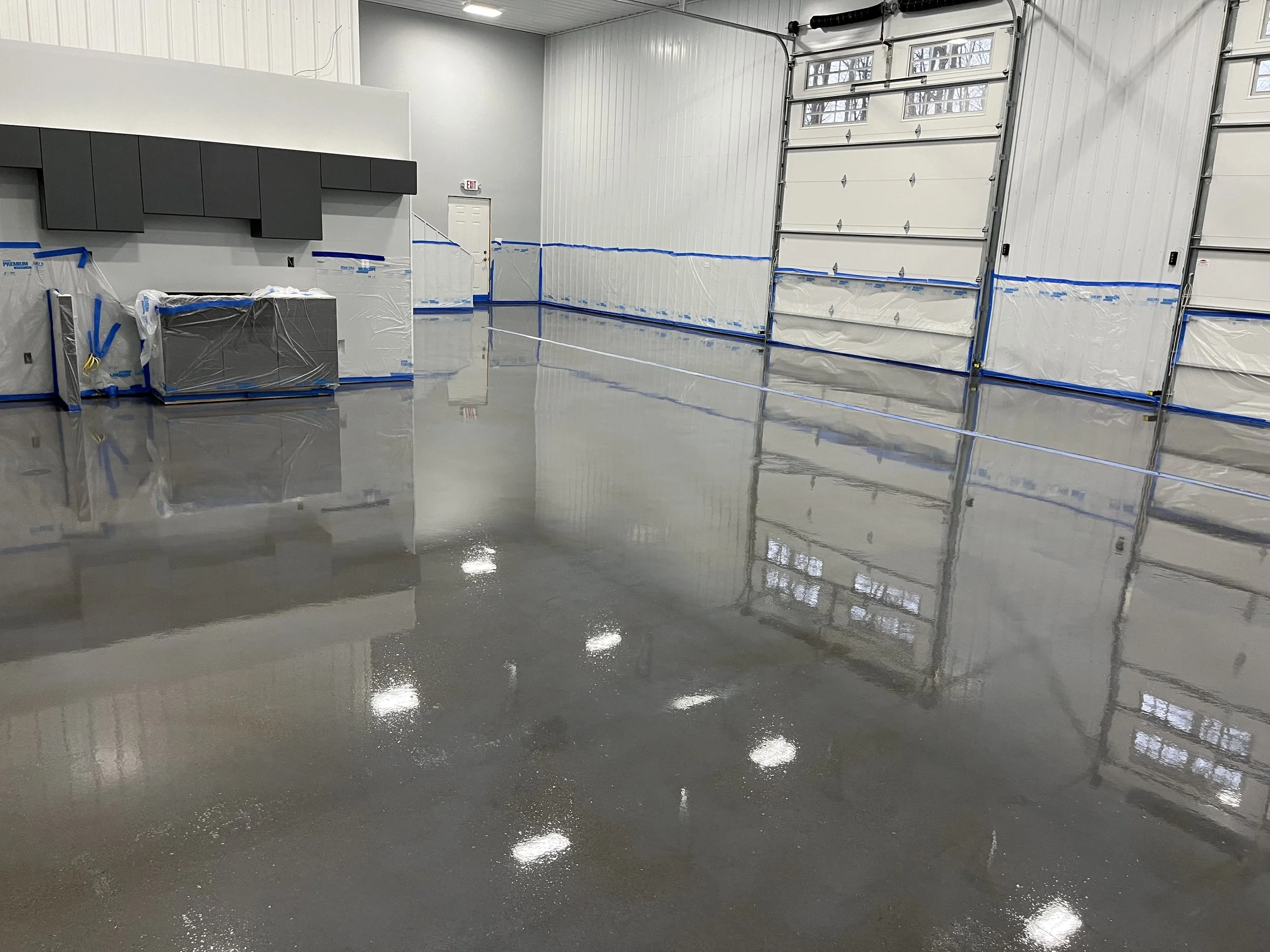 Empty garage with a polished concrete floor, white walls, and large garage doors; some construction materials covered with plastic and blue painter's tape along walls.