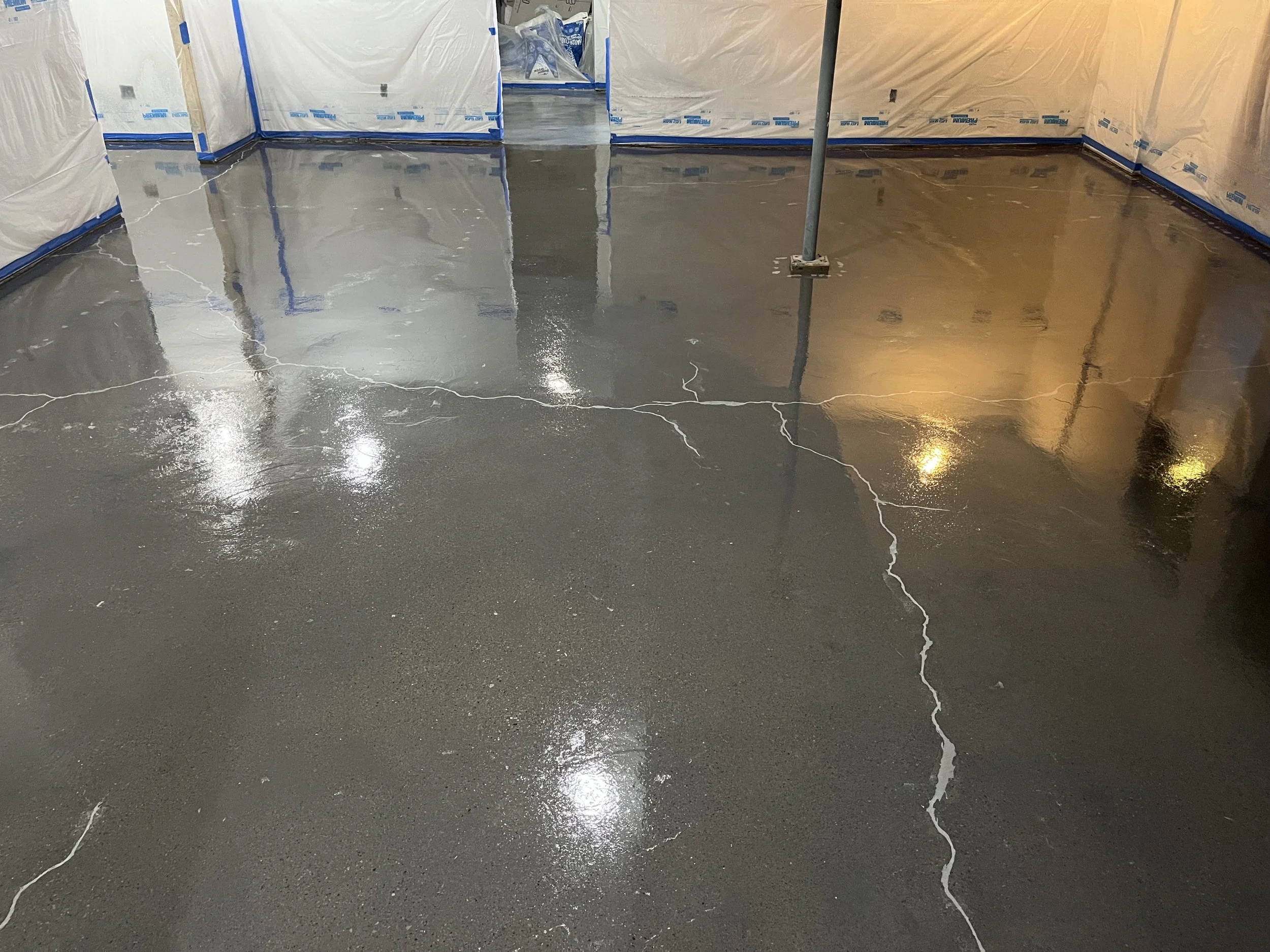 Concrete floor with visible cracks is wet and shiny, surrounded by temporary construction barriers covered with plastic sheeting.