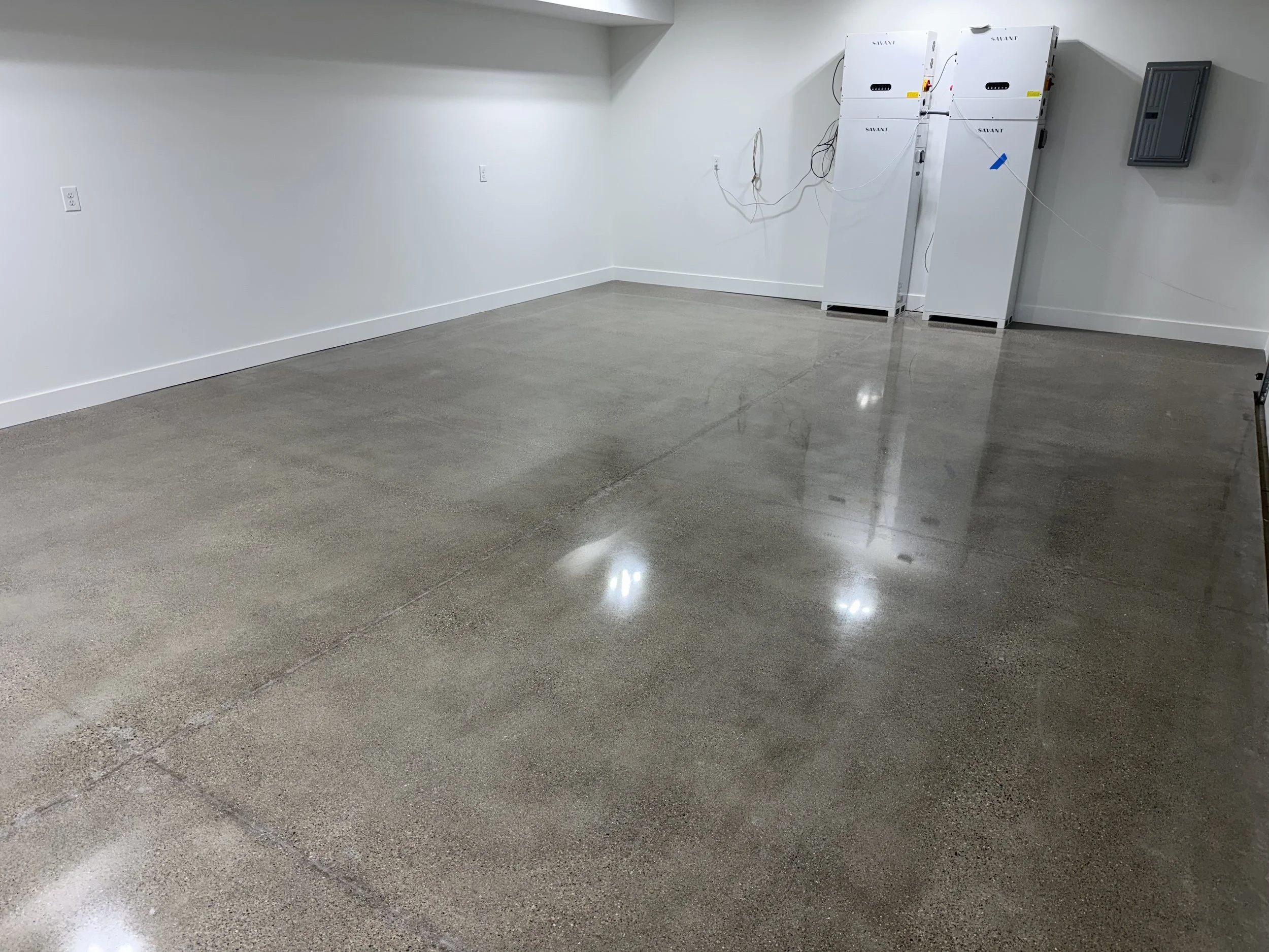 Empty room with white walls and a polished concrete floor. In the corner, there are three white machines connected with wires, and a gray electrical panel is mounted on the wall.