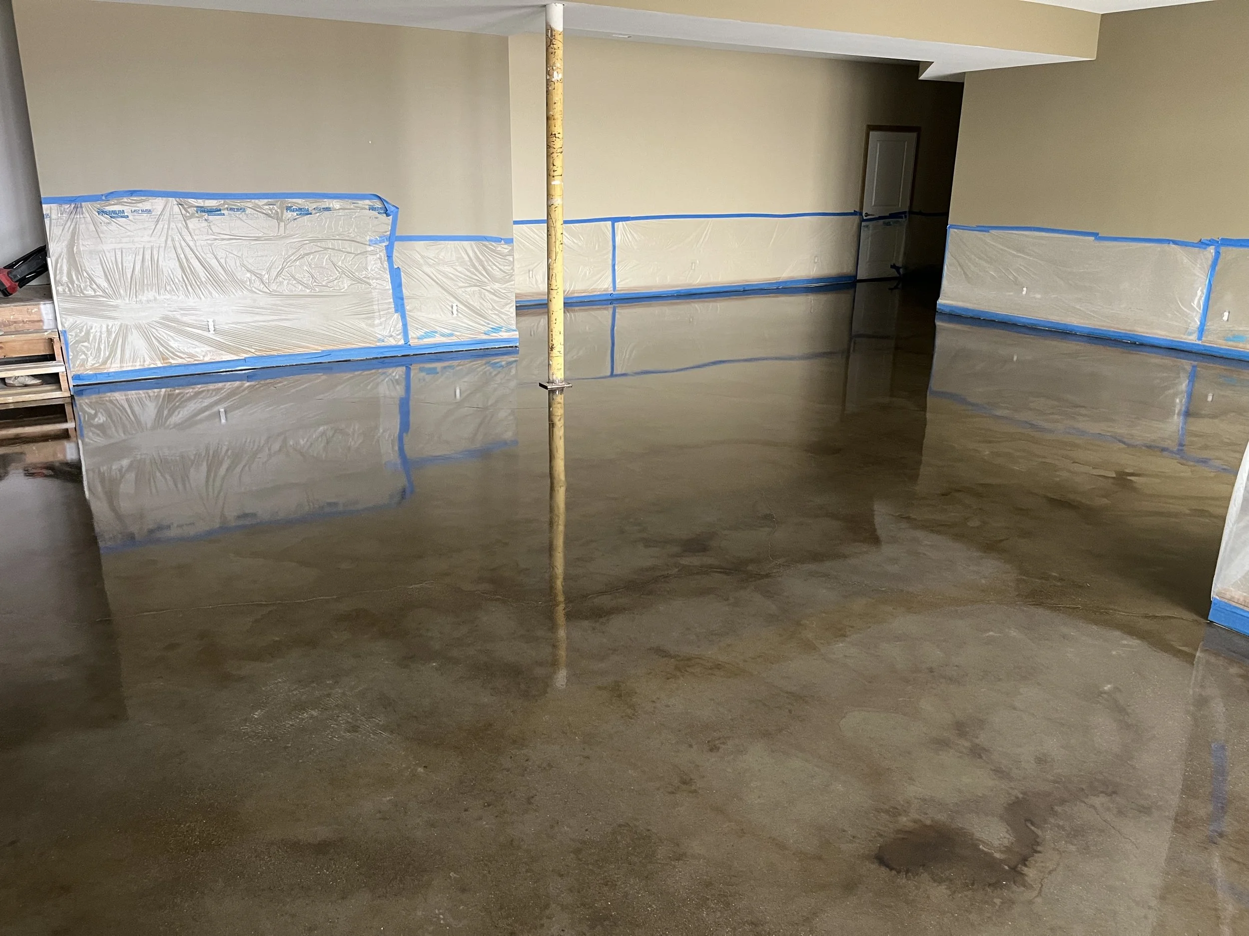 A room with a newly poured concrete floor that is still wet, reflecting the walls and ceiling. The walls are covered with plastic sheeting secured with blue painter's tape, indicating ongoing construction or renovation.