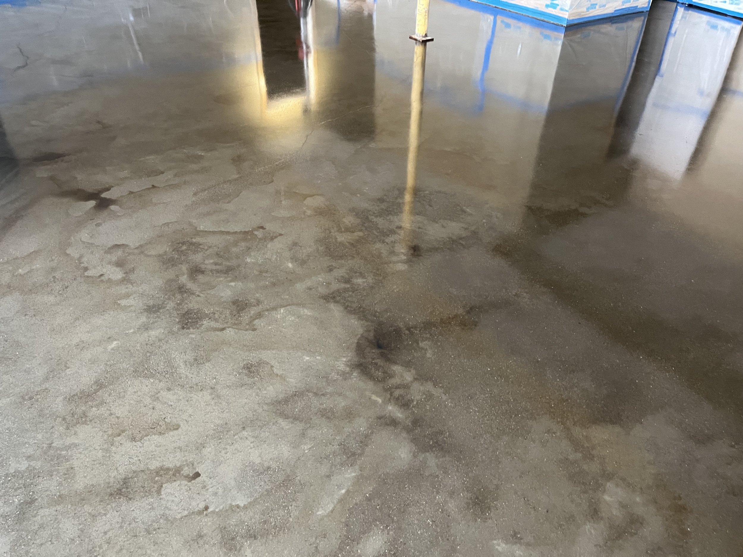 Concrete floor that is wet and patched with areas of dry concrete, in an indoor setting with construction equipment and partially covered walls.