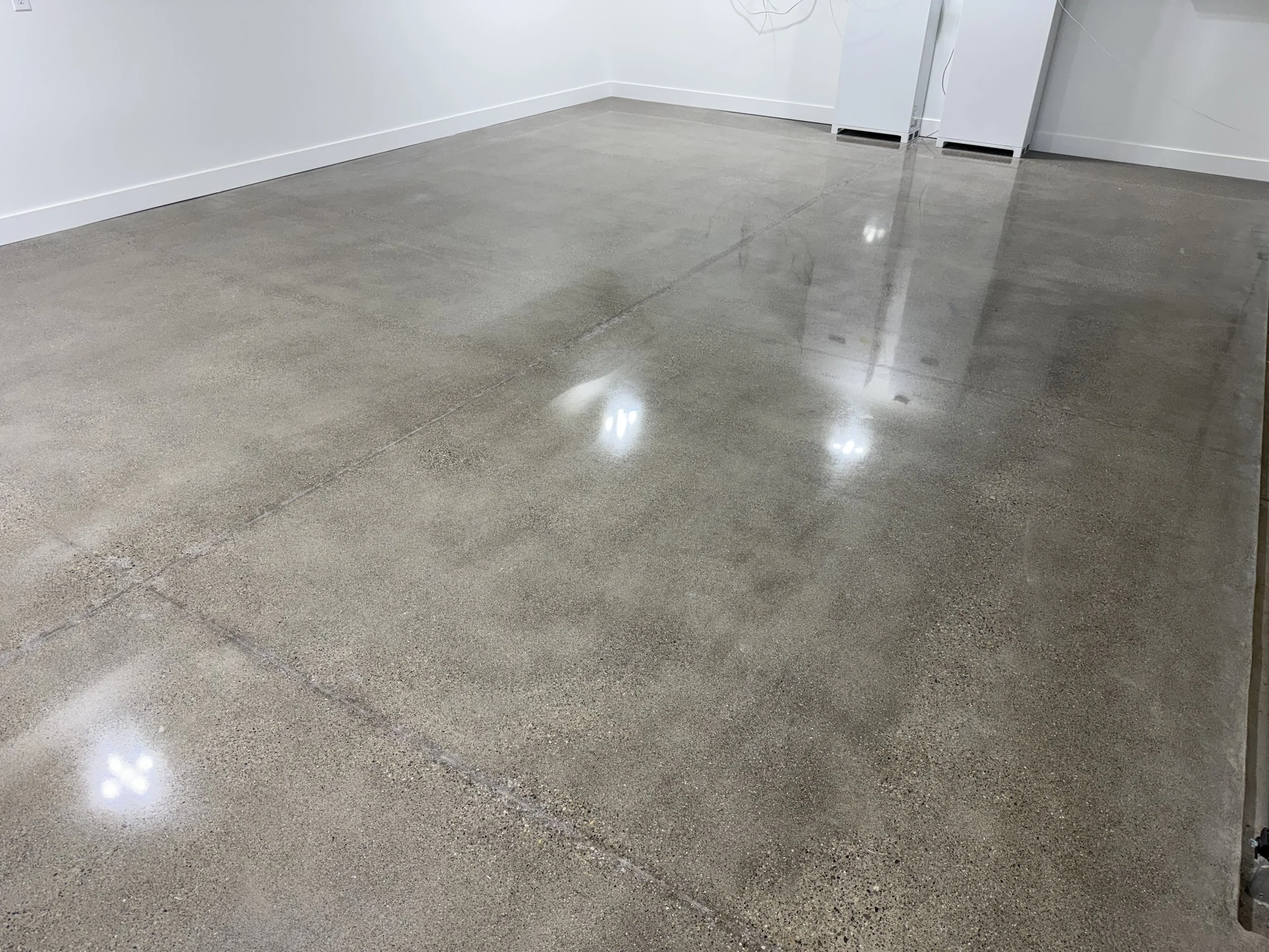 Polished concrete floor with a glossy finish in a room with white walls and baseboards, and visible furniture and appliance reflections.