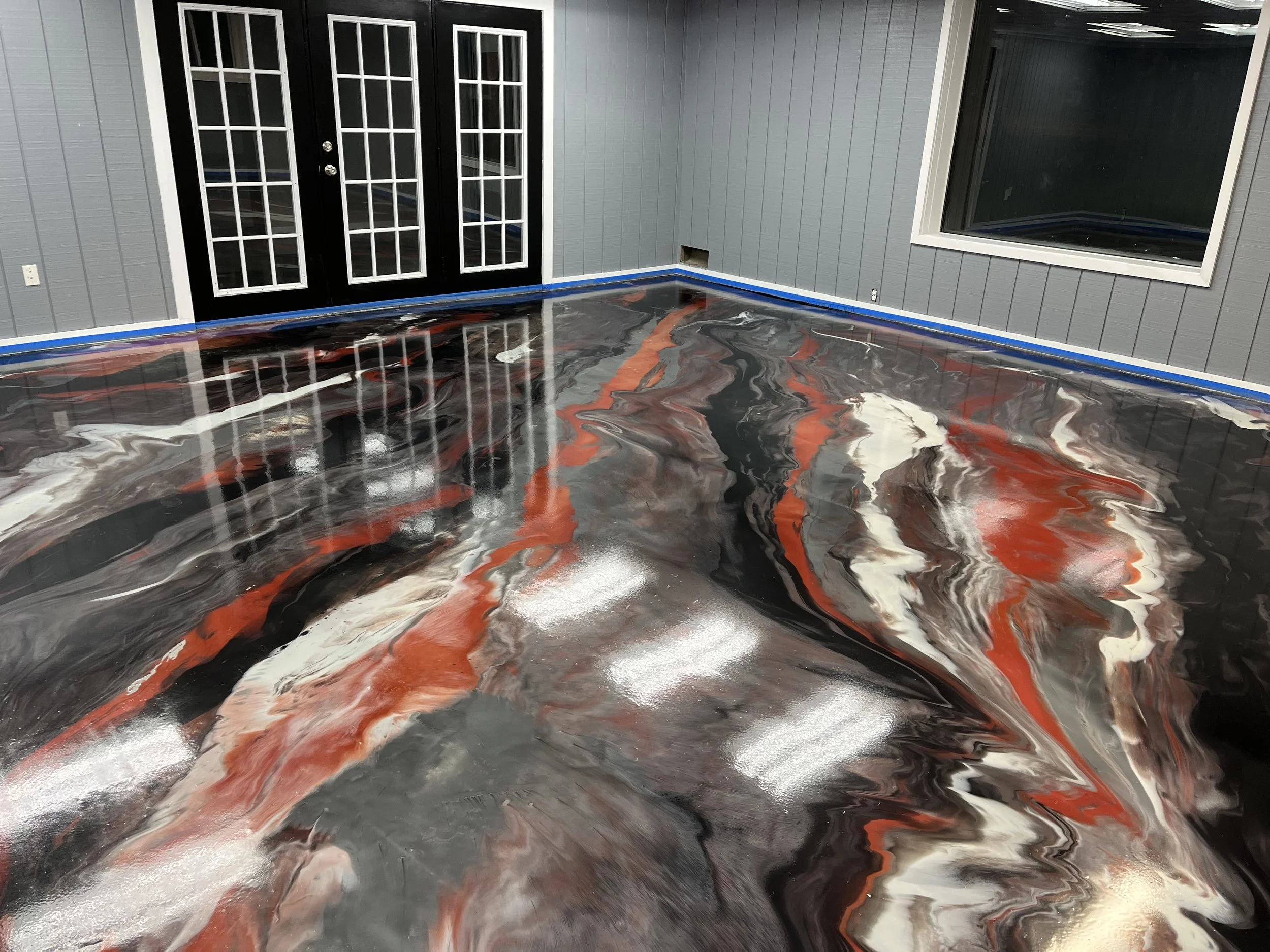 Interior of a room with a shiny, swirling black, white, and red epoxy flooring, gray walls, a black door with glass panels, and a large window.
