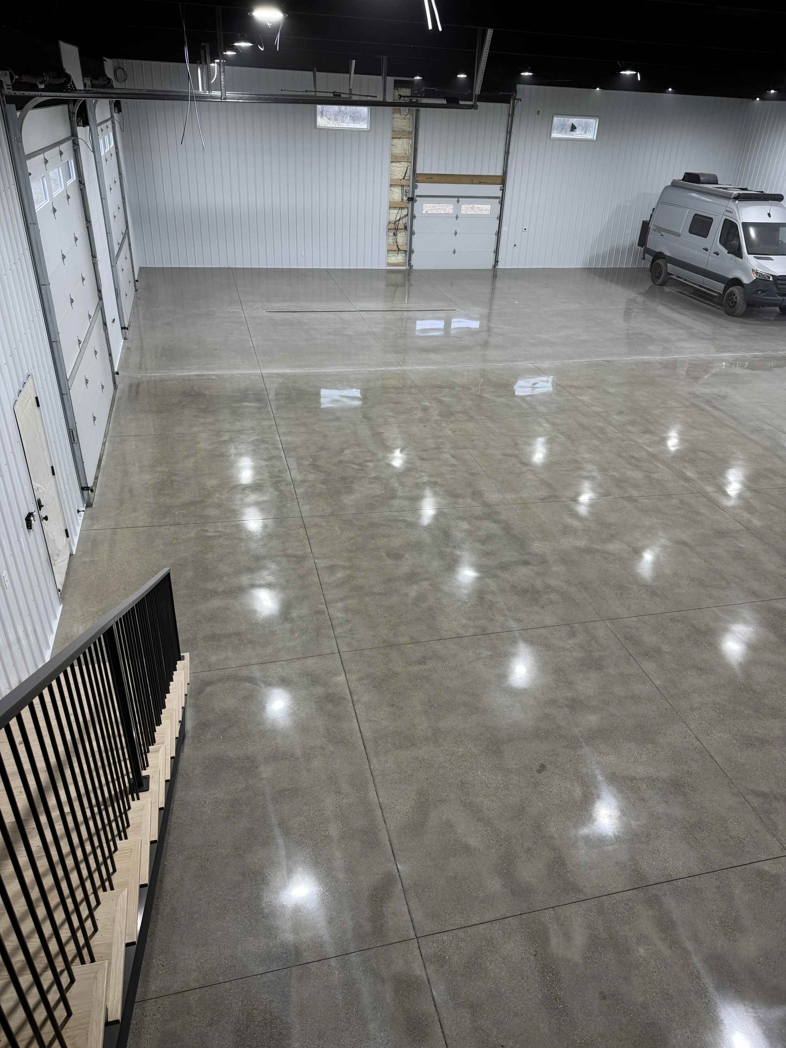 Empty commercial garage with polished concrete floor, metal walls, overhead doors, a van parked inside, and bright ceiling lights.