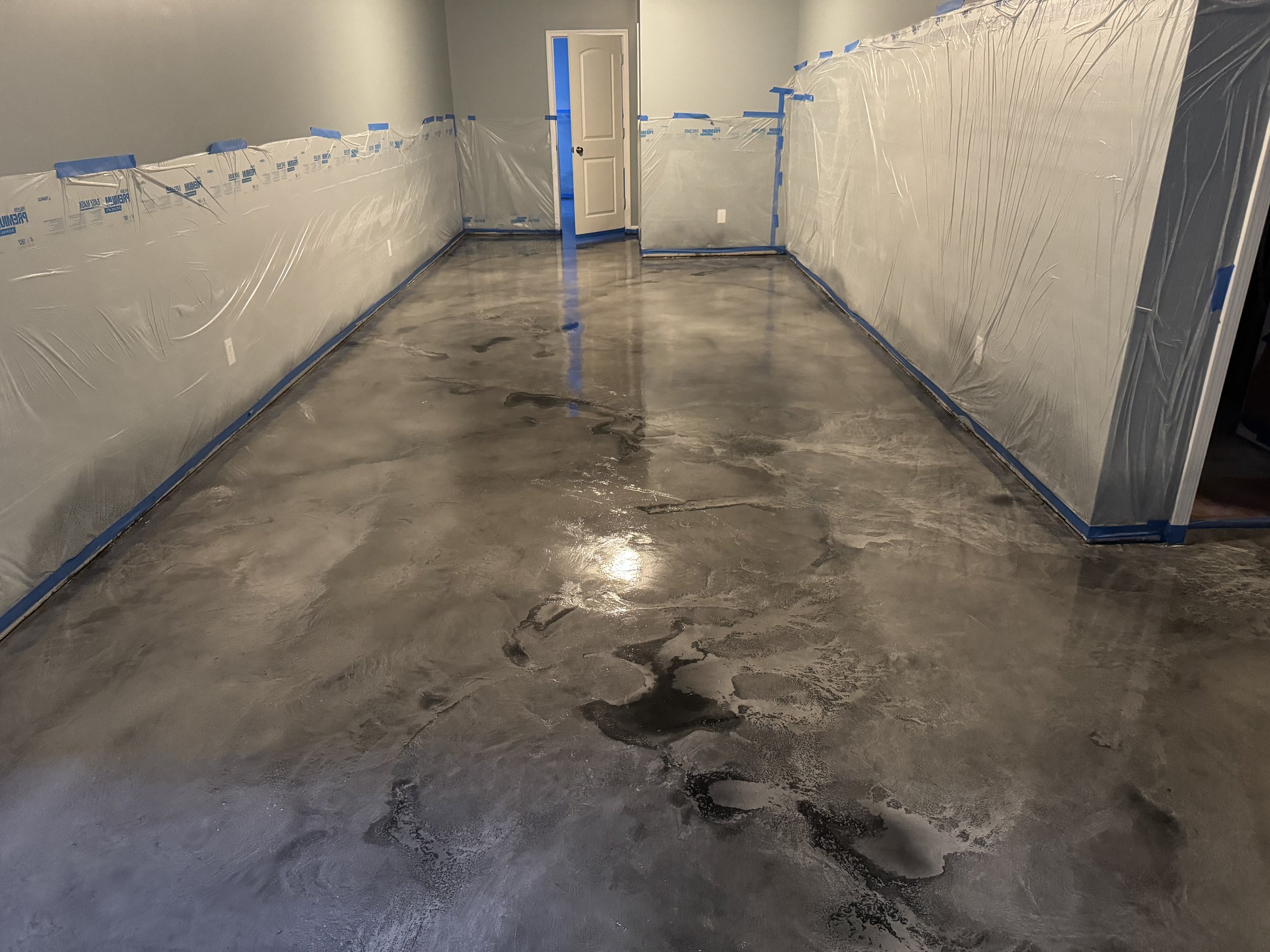 A room with a freshly poured and sealed concrete floor. The walls are partially covered with plastic sheeting taped along the edges, indicating ongoing construction or renovation.
