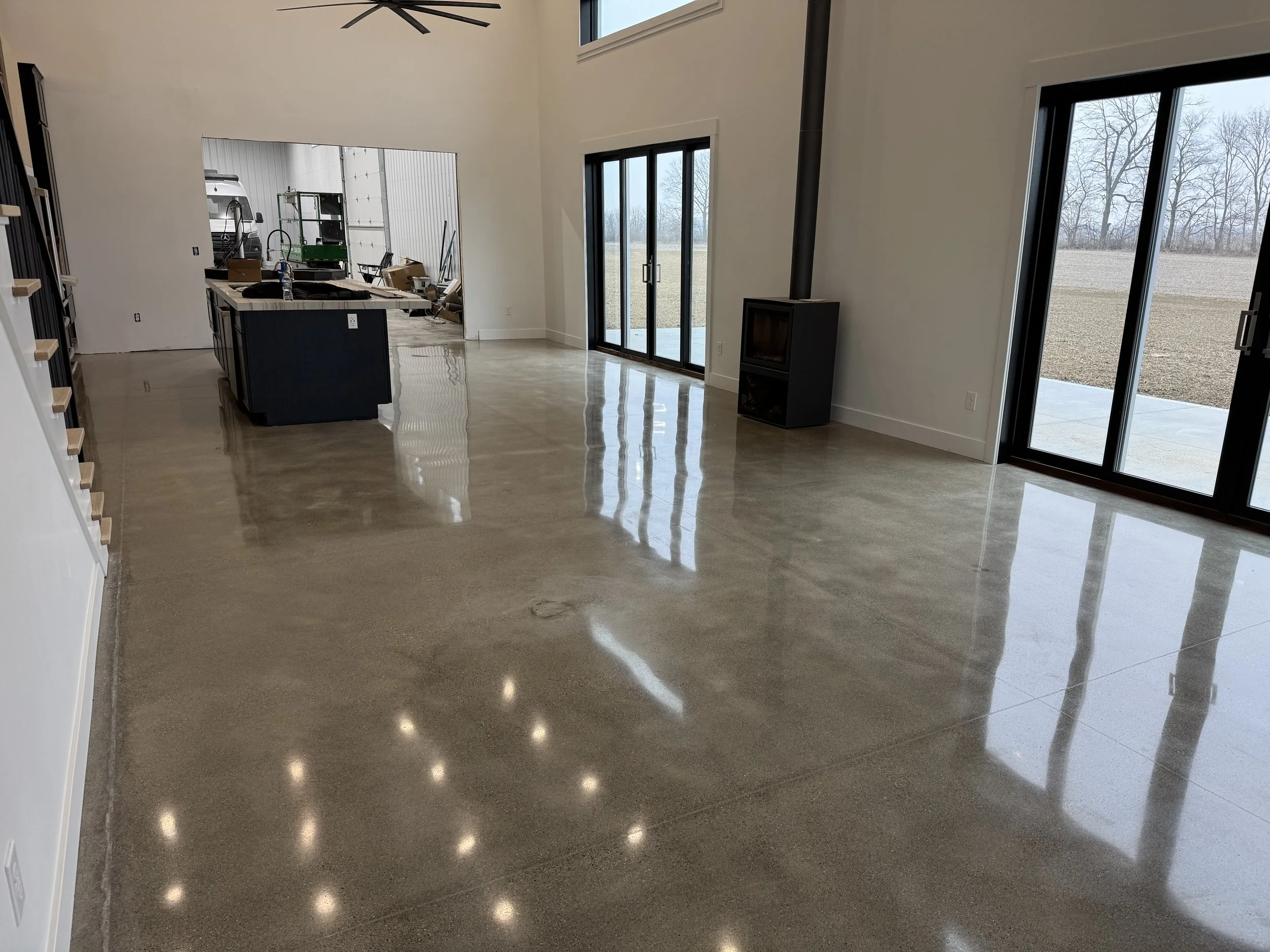 Empty modern living room with polished concrete floors, large glass sliding doors, a wood stove, and an open kitchen area in the background.