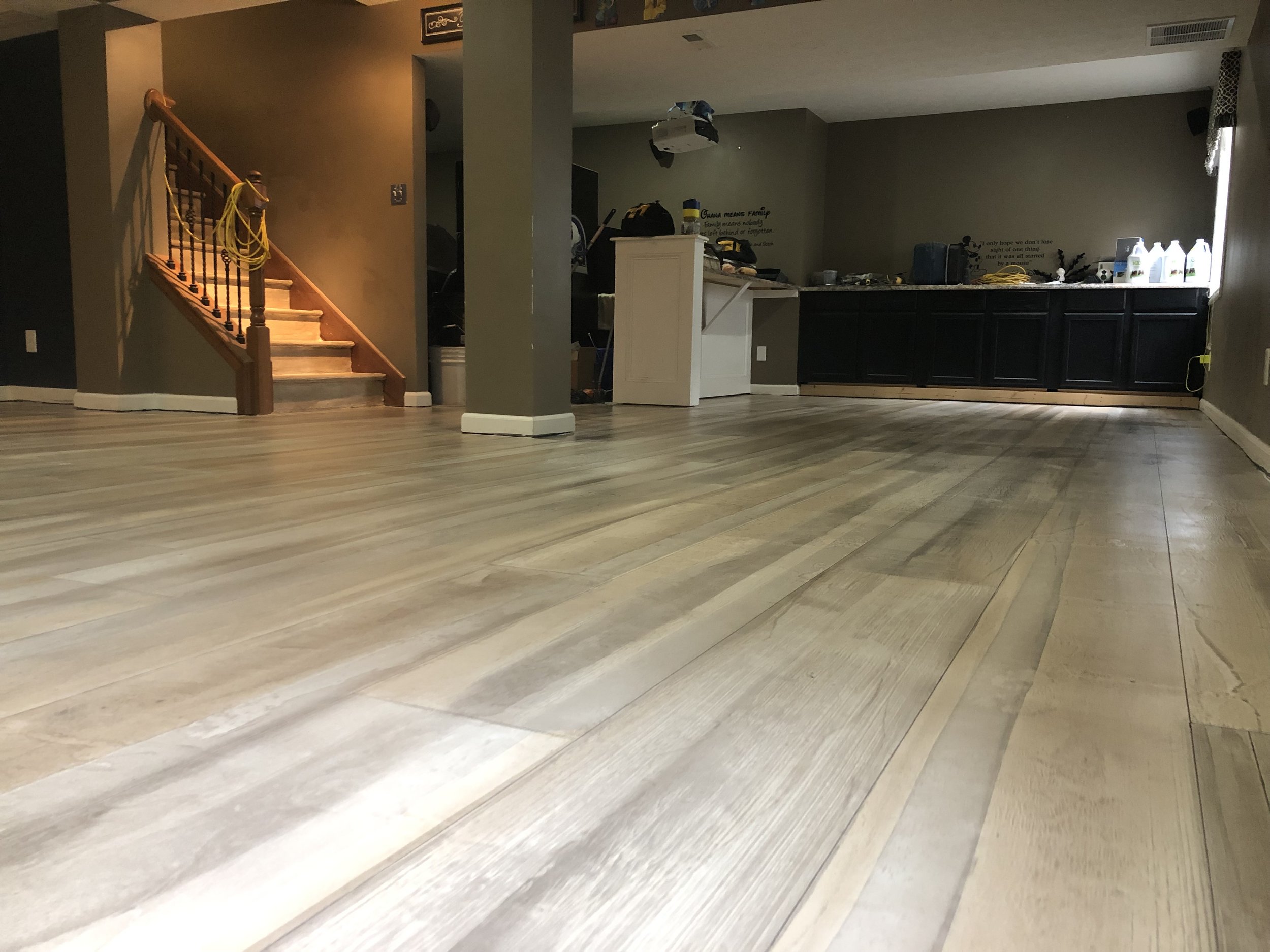 Empty room with newly installed light-colored wooden flooring, a staircase with wooden handrail on the left, dark cabinetry and supplies on the back wall, and a window with a curtain on the right.