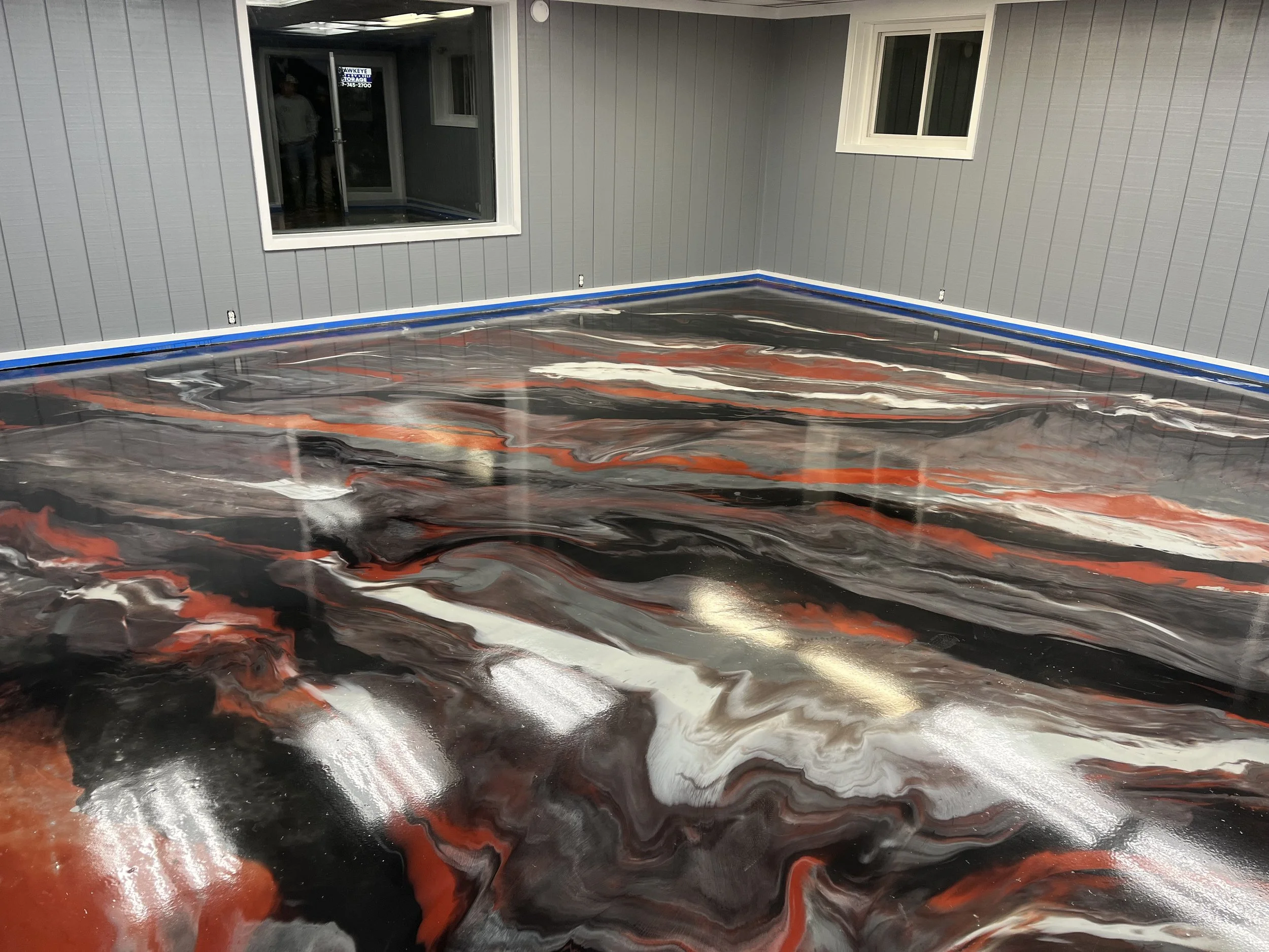 A newly poured concrete floor in a room, with swirling black, white, and red epoxy finish, adjacent gray paneled walls, and windows with white frames.