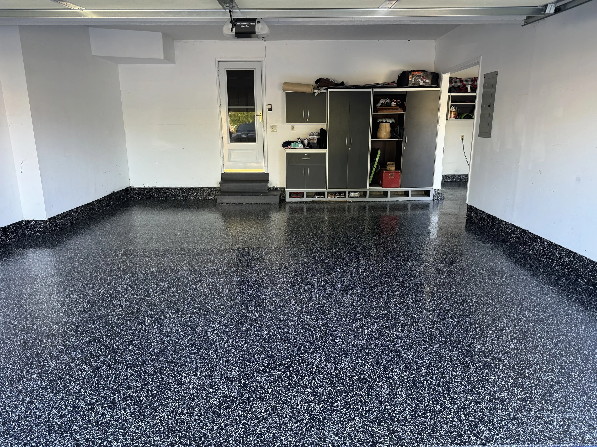 Empty garage with black speckled epoxy flooring, white walls, and a white door leading outside. There are shelves with tools and storage items on the back wall.