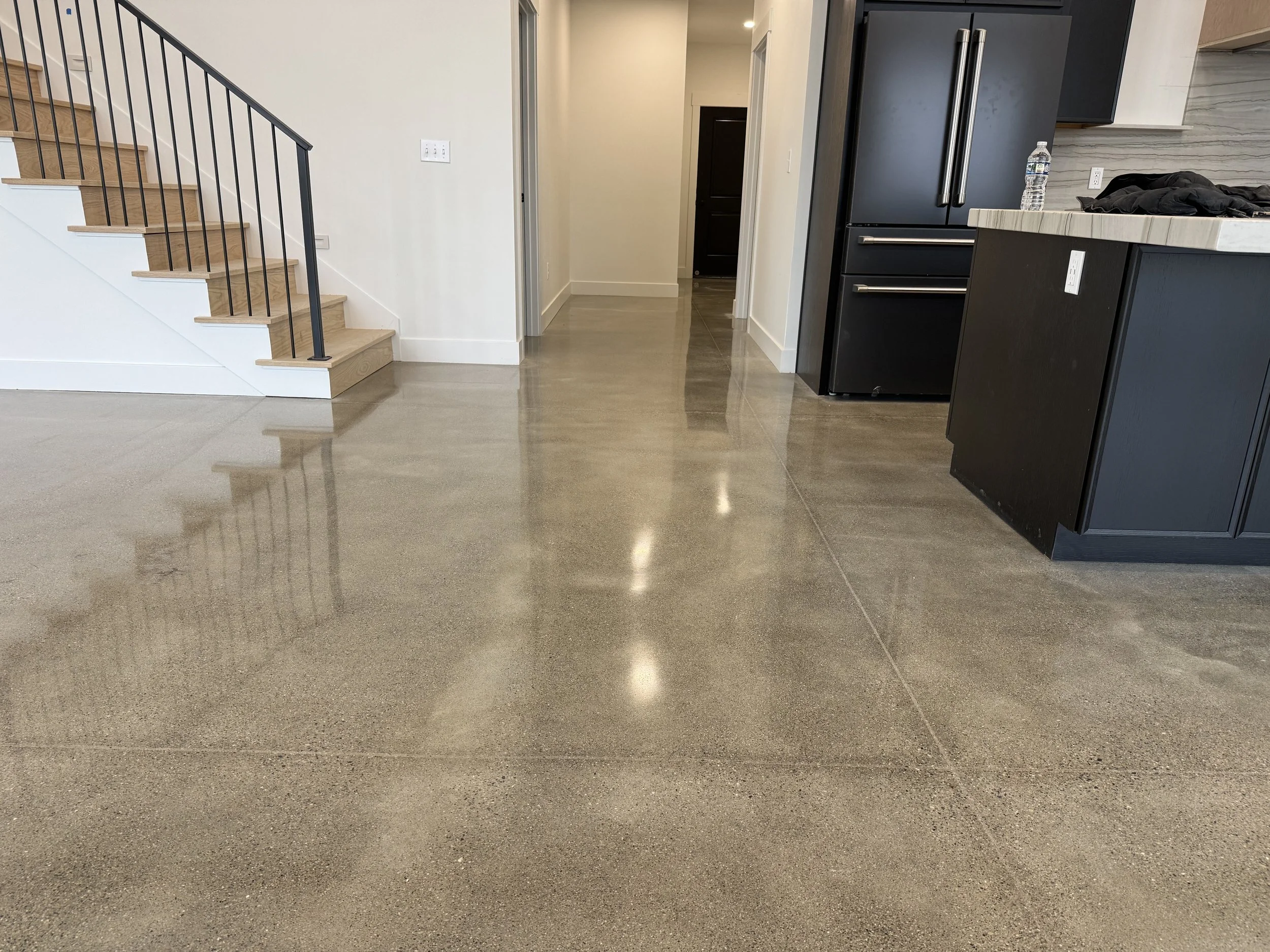 Polished concrete floor in a modern kitchen with black cabinets, a black refrigerator, and a staircase with wooden steps and black railing.